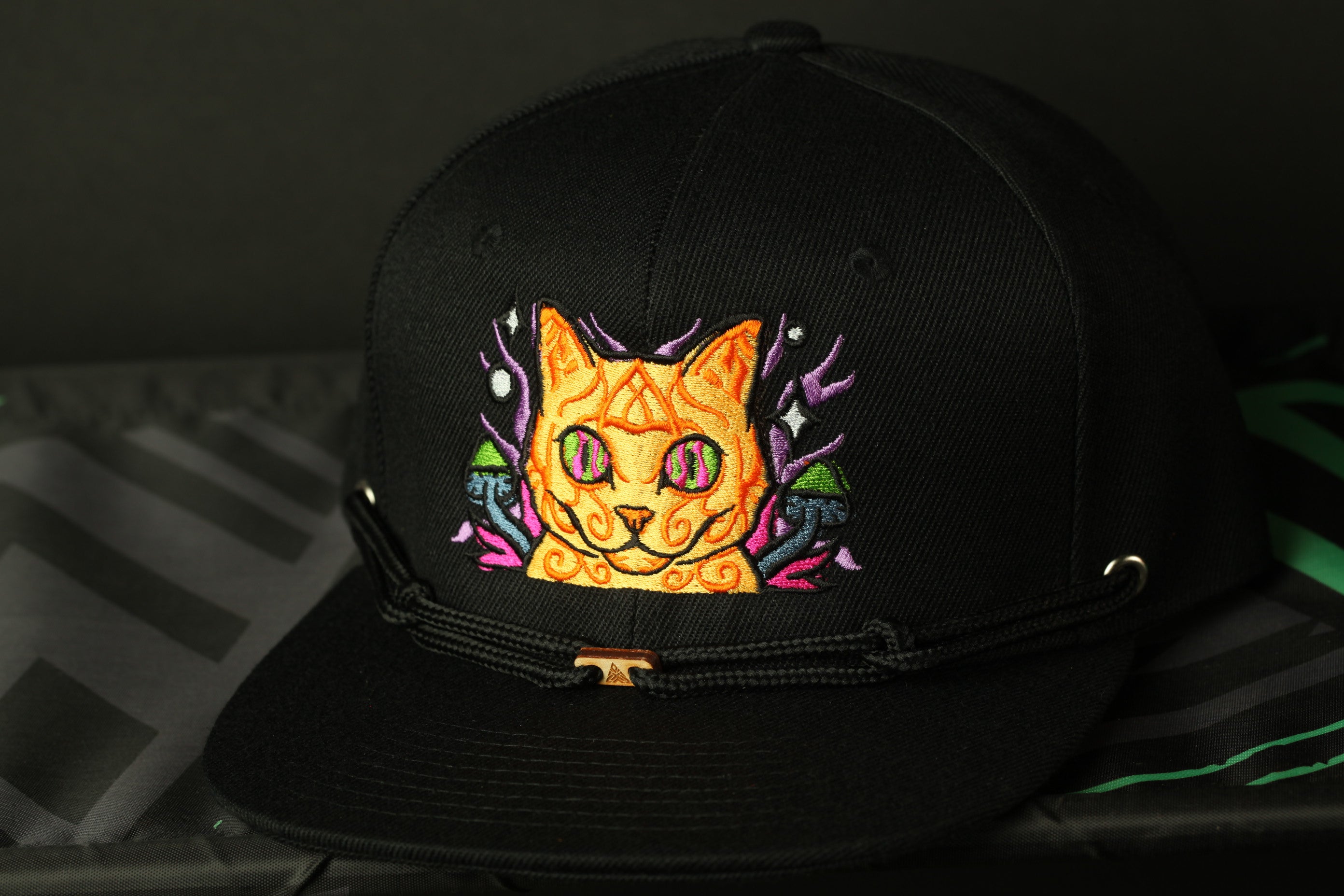 A black baseball cap with a colorful cat embroidered on it, featuring a yellow cat with a purple and pink design around its face.