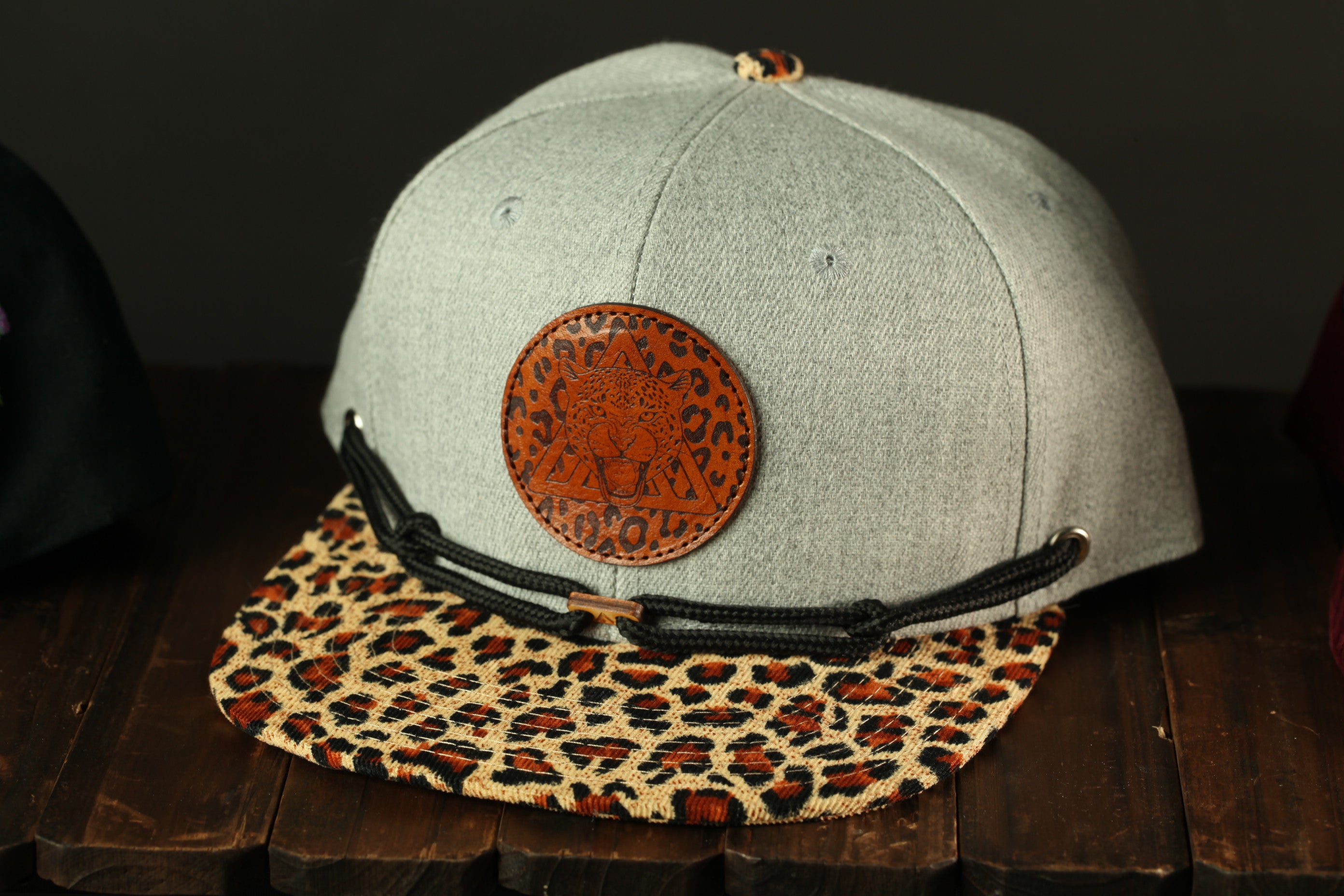 A gray baseball cap with a leopard print pattern and a circular logo on the front, placed on a wooden surface.