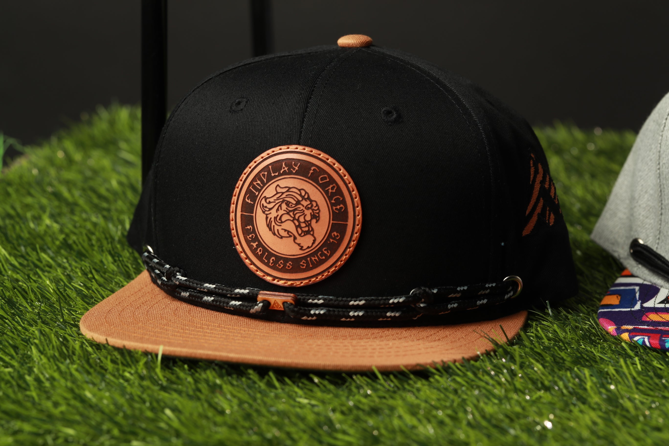 A black baseball cap with a brown leather patch and a black and white braided strap, placed on a grassy surface.