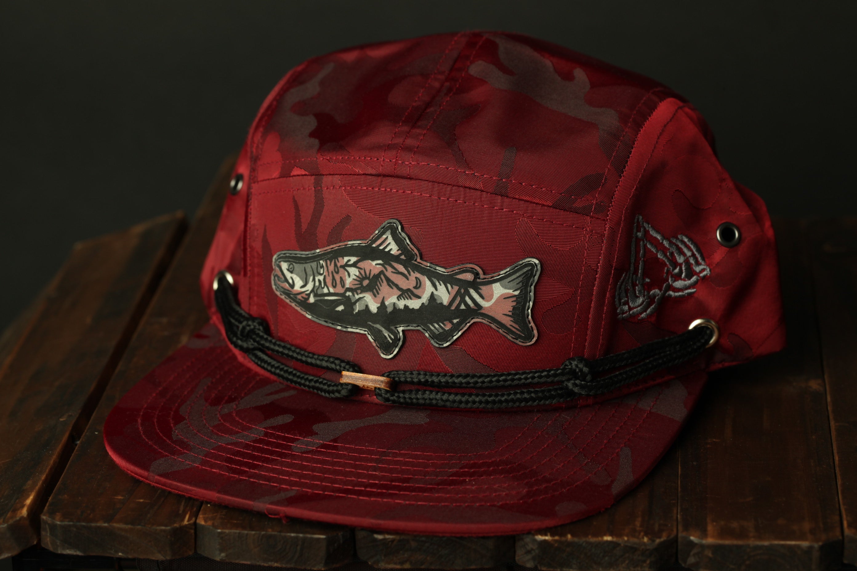 A red camouflage baseball cap with a fish patch on the front, sitting on a wooden surface.