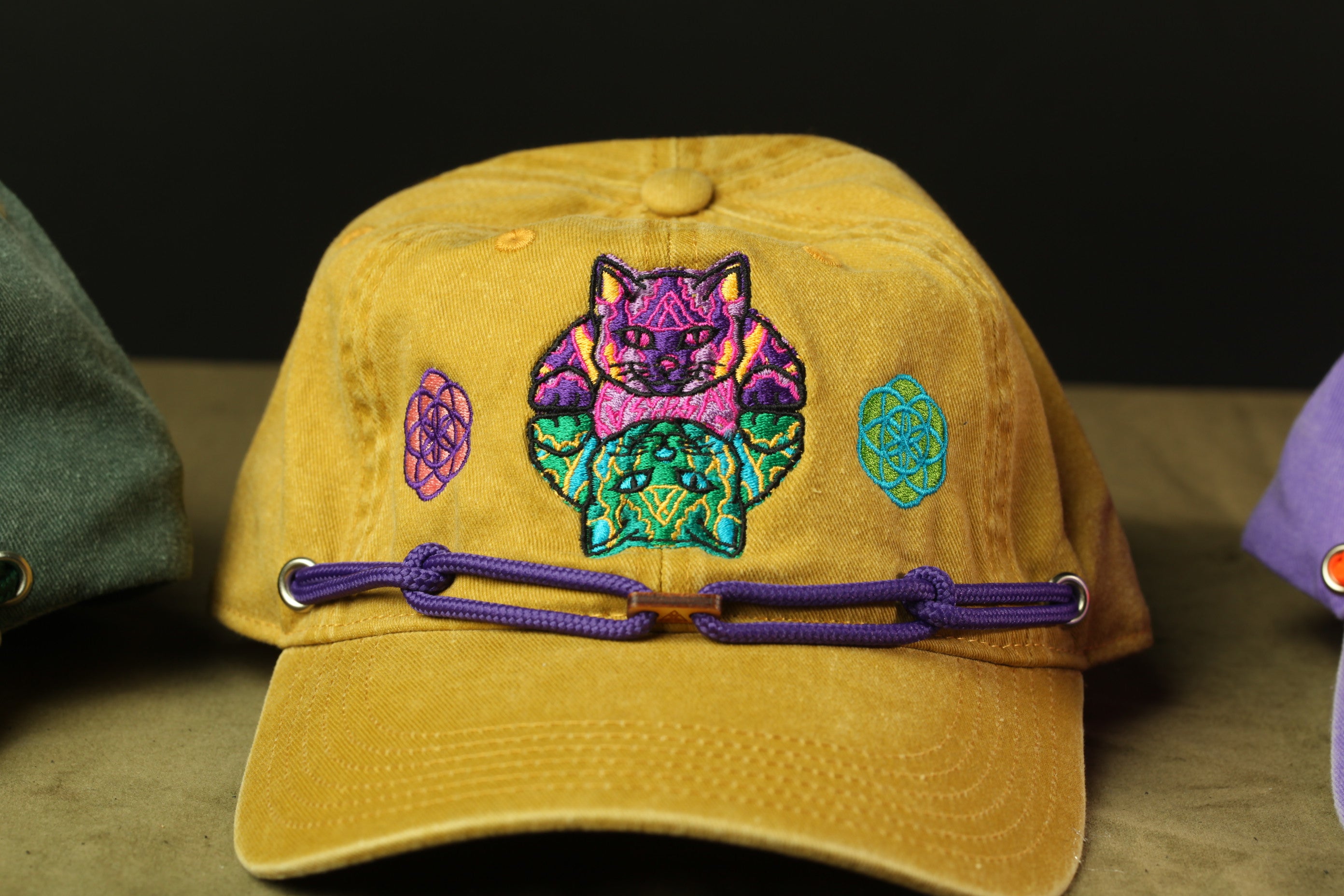 A yellow baseball cap with a colorful cat embroidered on it, featuring a purple ribbon around the brim.