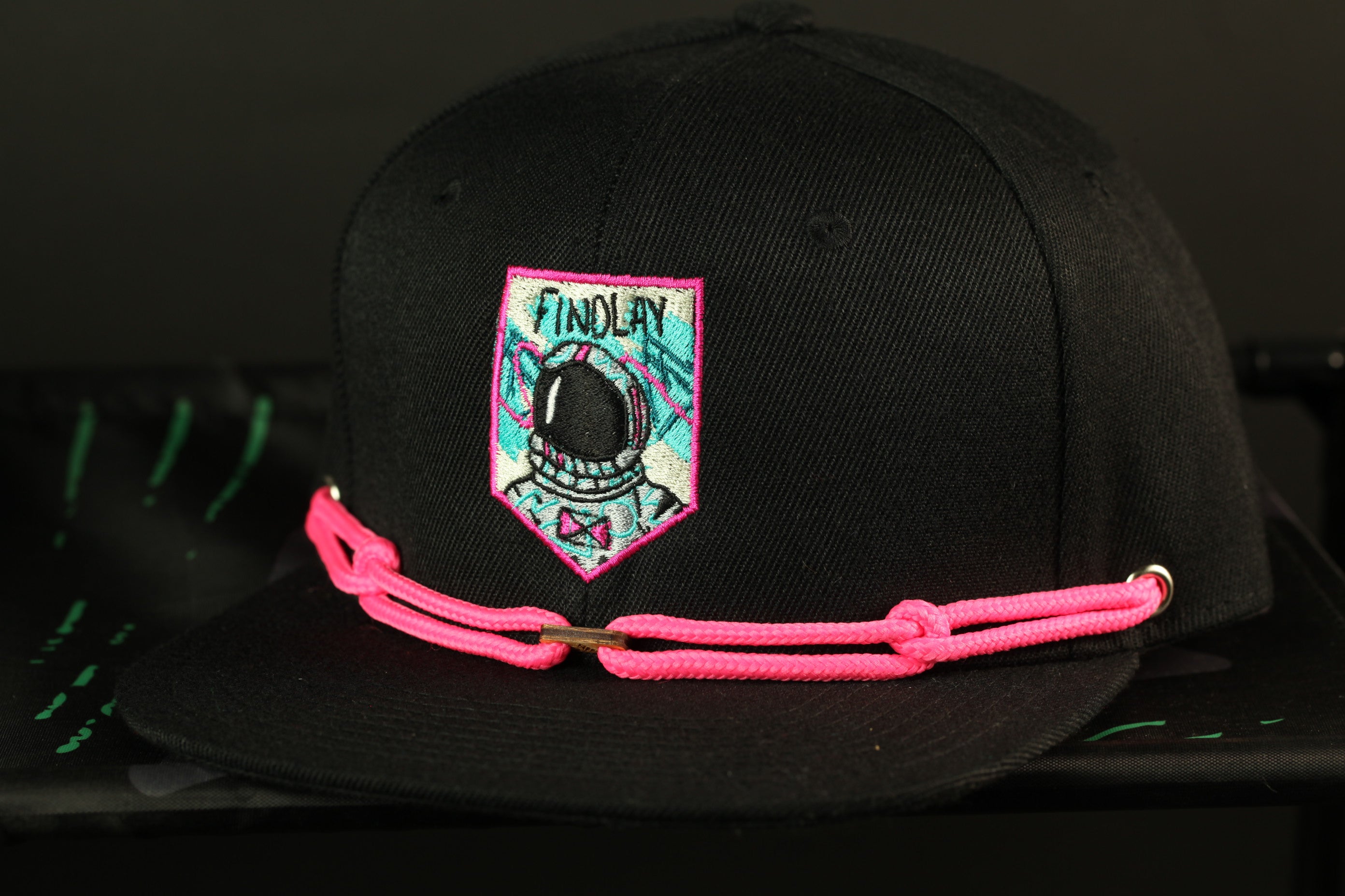 A black baseball cap with a pink chain strap and a patch on the front, featuring a graphic of a person wearing a helmet.