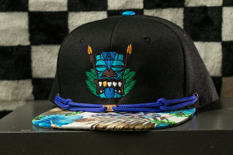 A black baseball cap with a colorful, embroidered design on the front, featuring a blue and green mask-like figure with a tiki-style headdress.