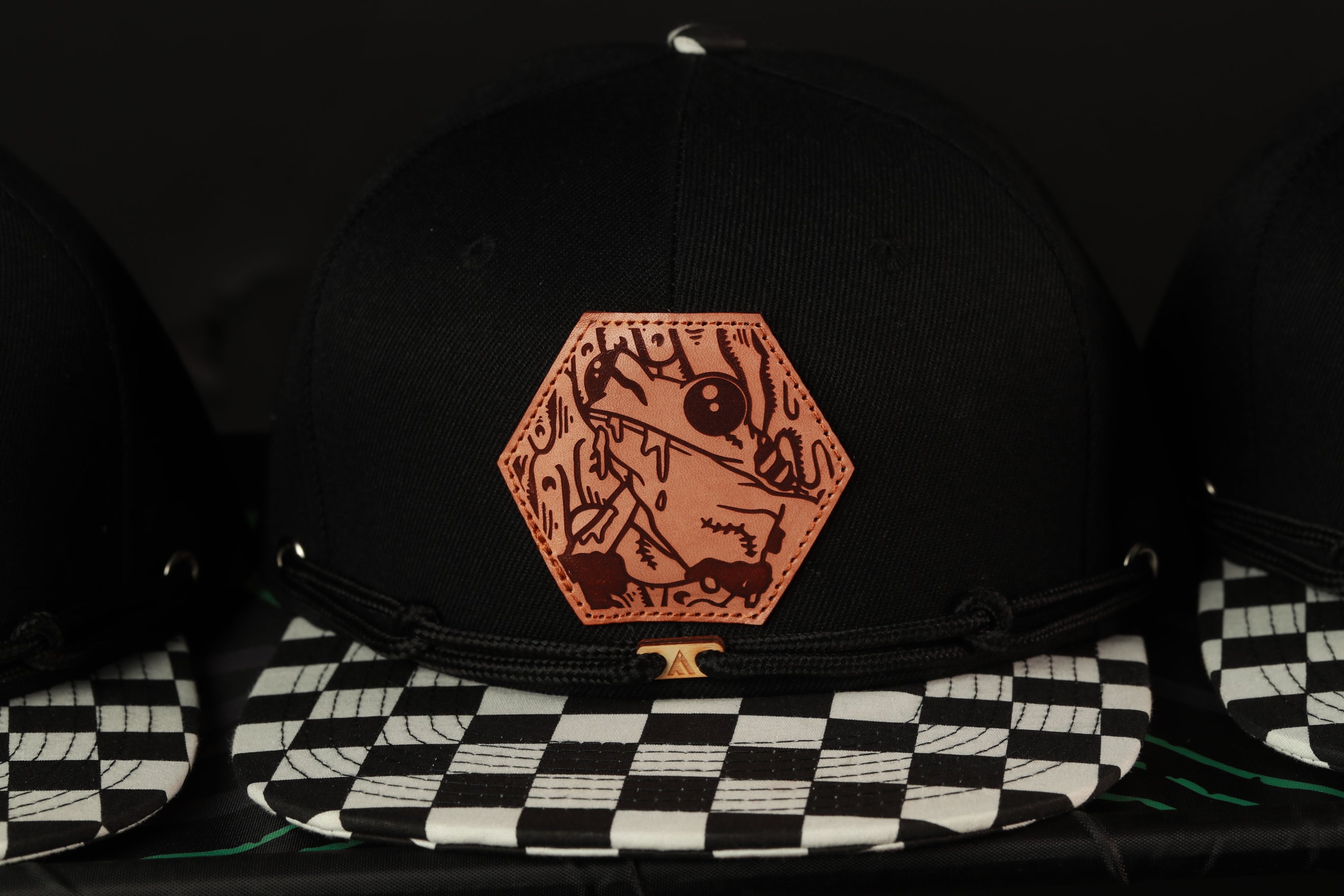 A black baseball cap with a distinctive orange hexagonal patch featuring a cartoon character, placed on a black and white checkered surface.