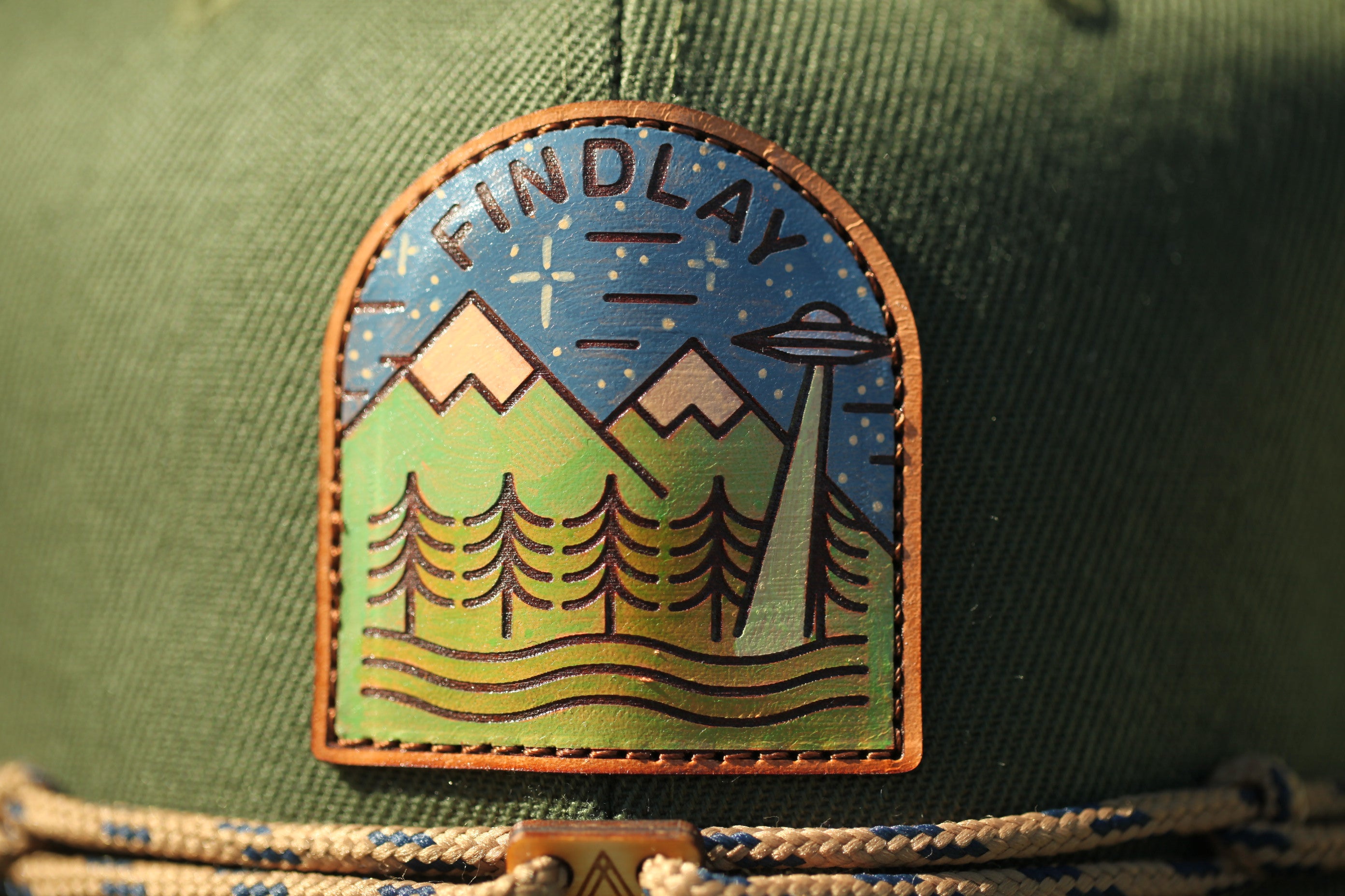 A close-up of a green hat with a patch featuring a mountain landscape, trees, and an alien spacecraft.