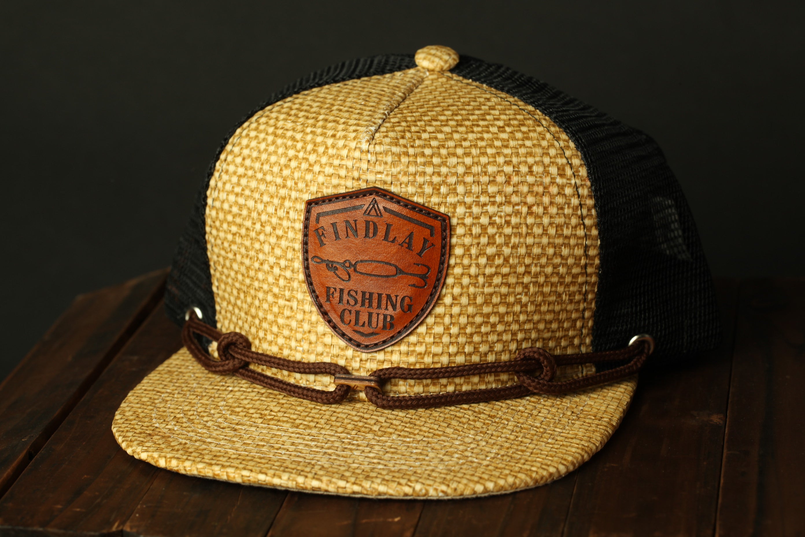 A straw hat with a black mesh back and a brown leather patch on the front, displaying the text "FINDLAY FISHING CLUB".