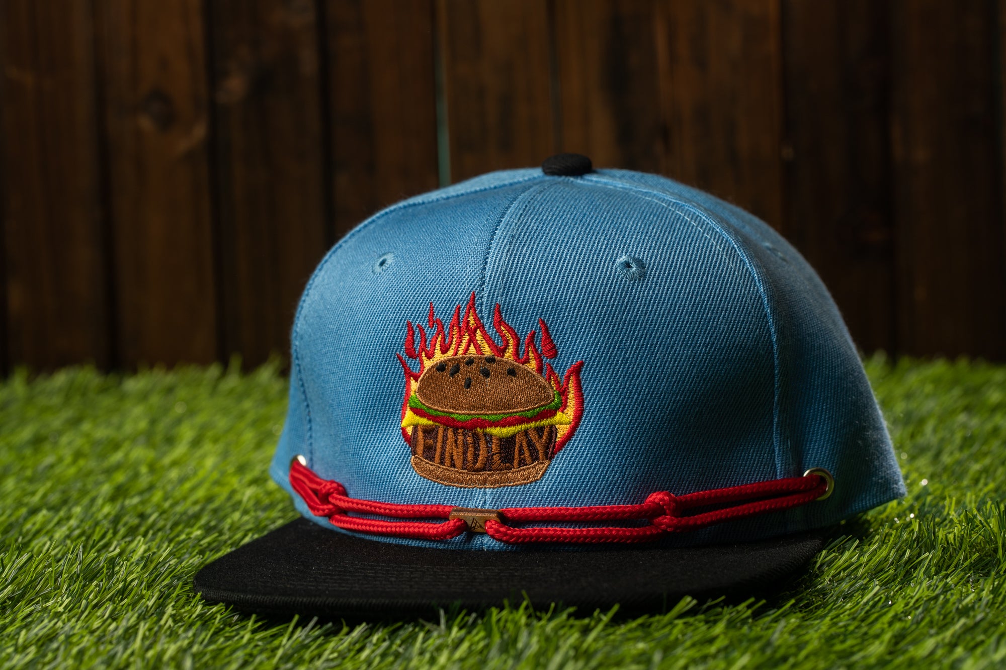 A blue baseball cap with a red string and a graphic of a hamburger on it, placed on a grassy surface.