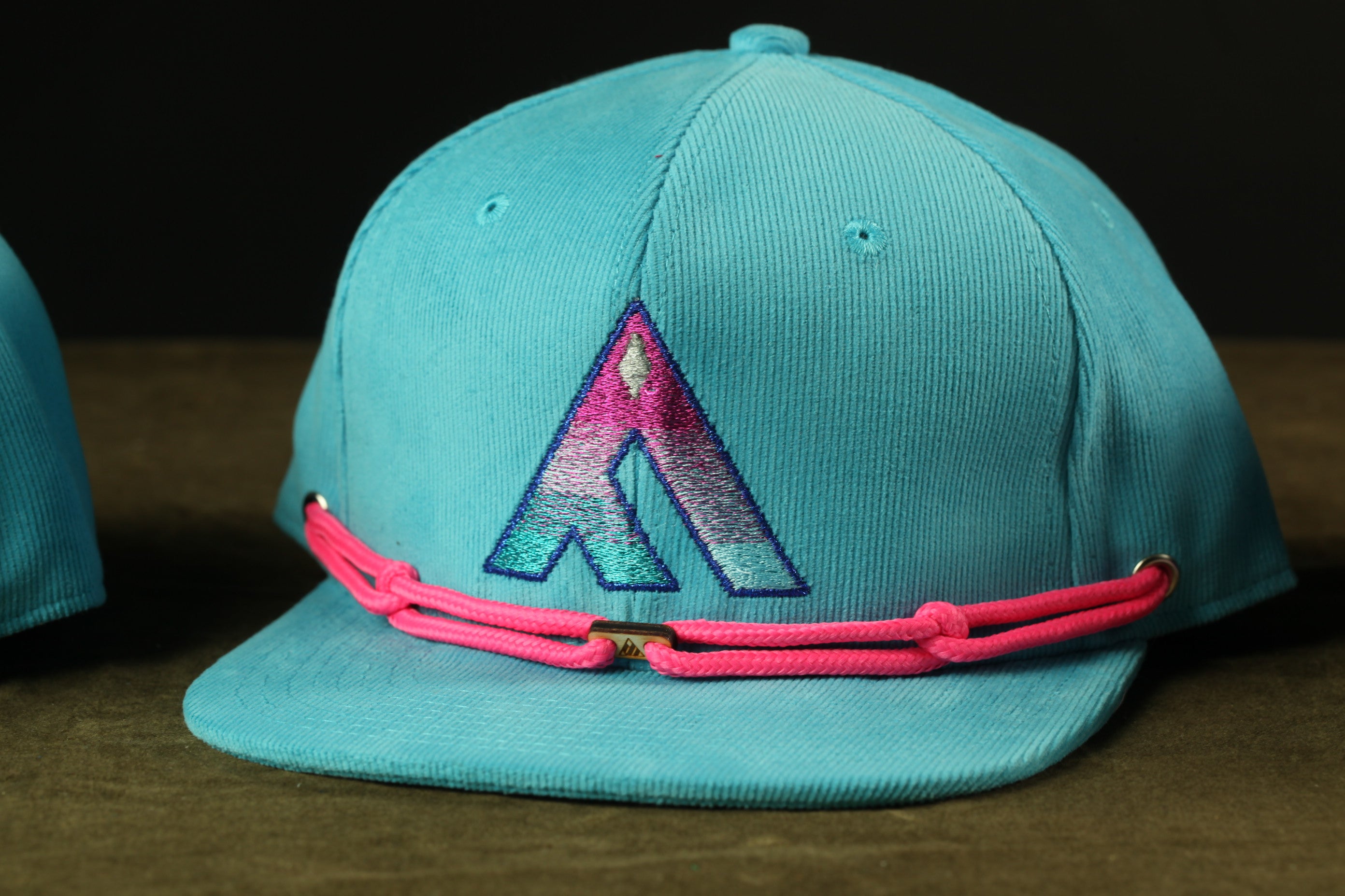 A turquoise baseball cap with a pink braided strap and a large embroidered logo on the front.