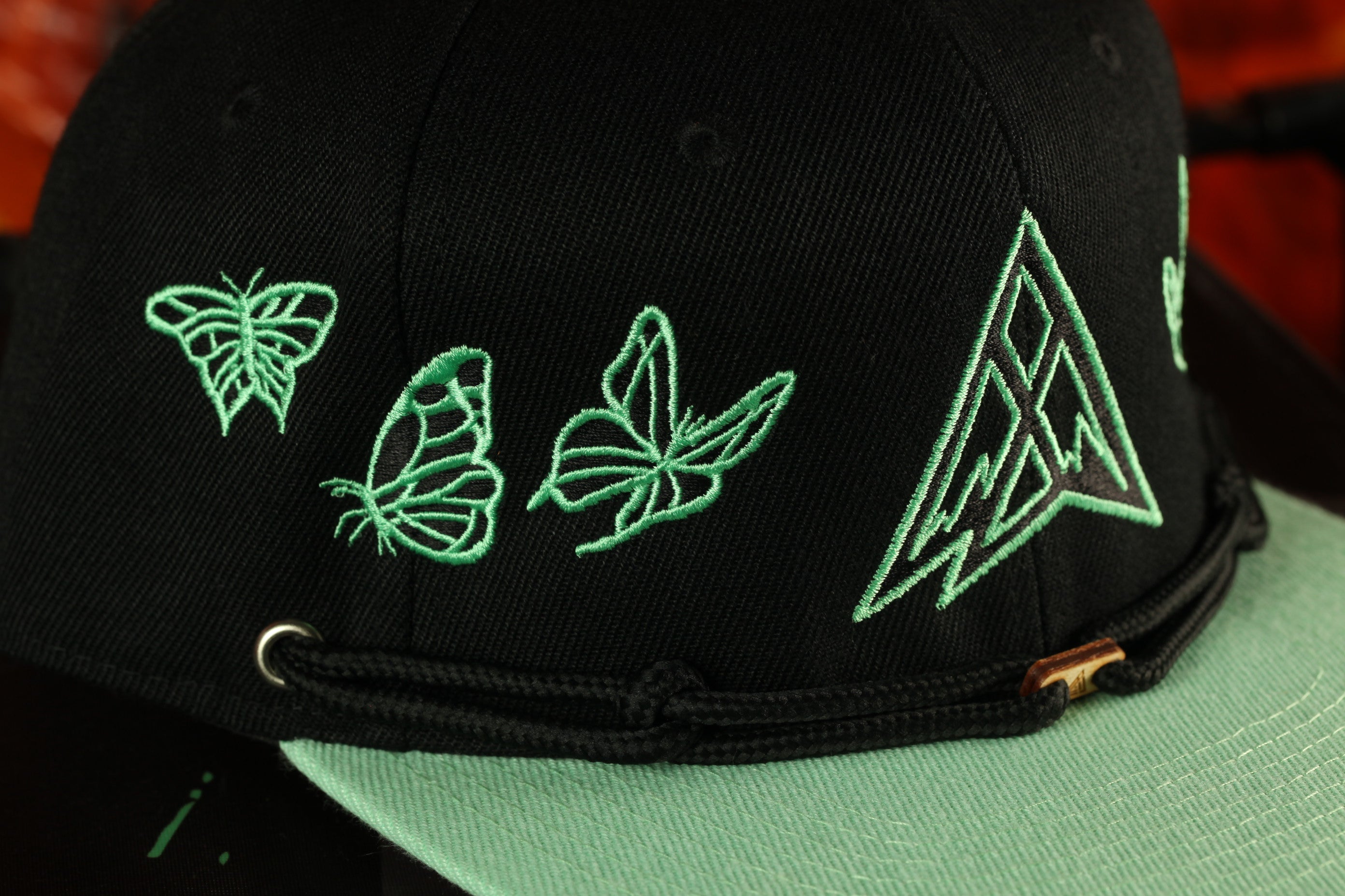 A close-up view of a black baseball cap with green embroidered designs, including butterflies and a geometric pattern.