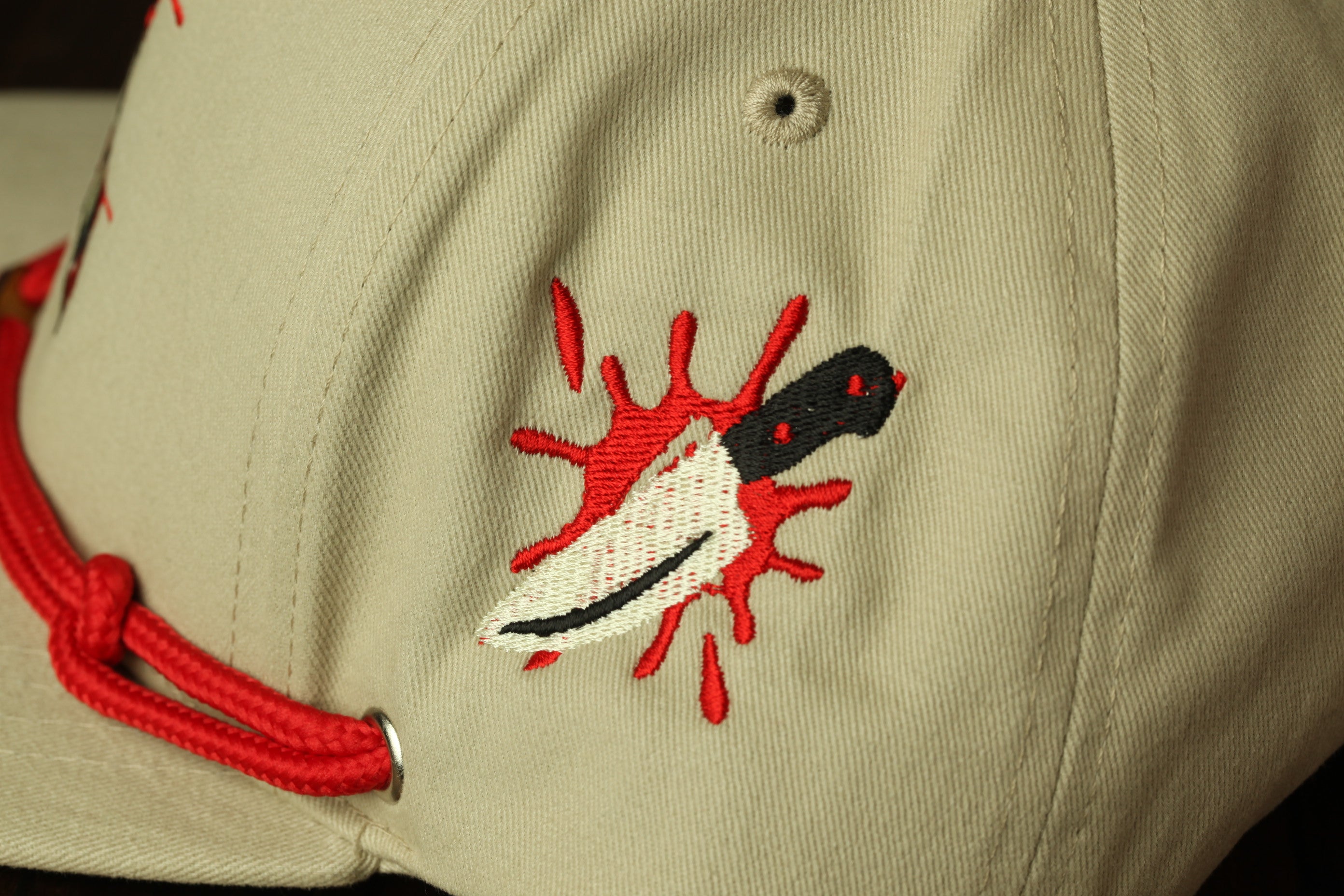 A close-up view of a beige baseball cap with a red string and a black and white embroidered image of a knife with a red splatter on the front.