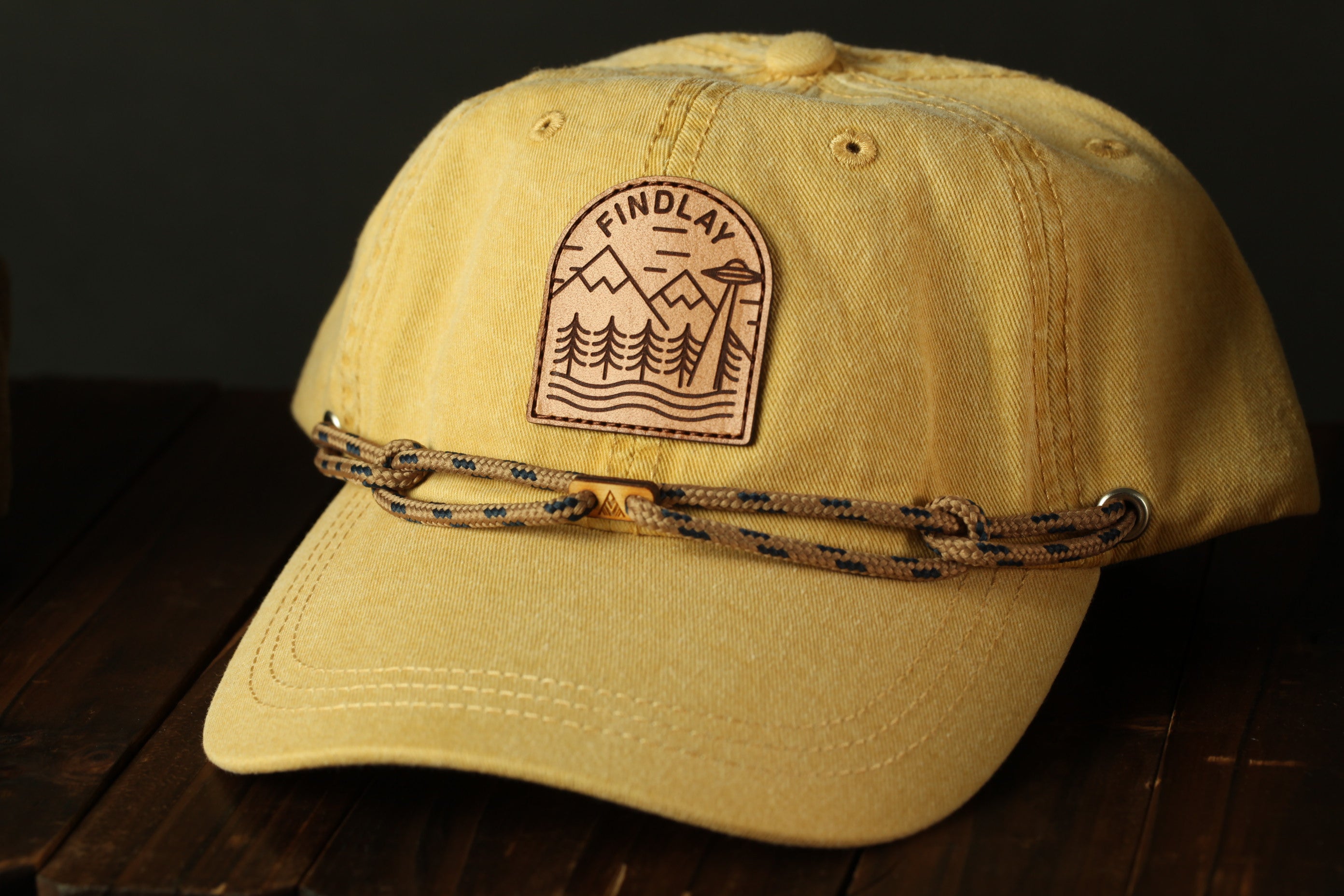 A yellow baseball cap with a leather patch on the front, featuring a mountain landscape design.