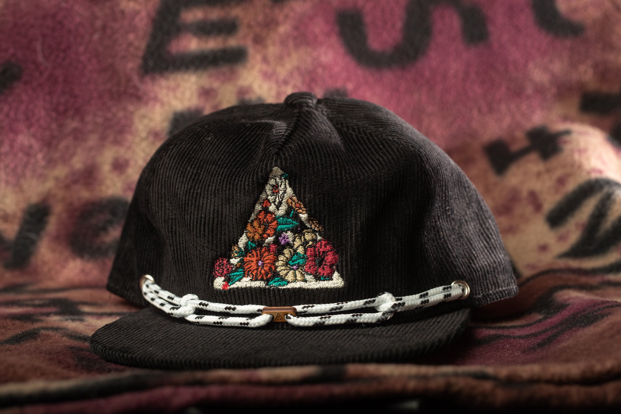 A black baseball cap with a floral embroidered triangle design on the front, resting on a pink and brown patterned fabric.