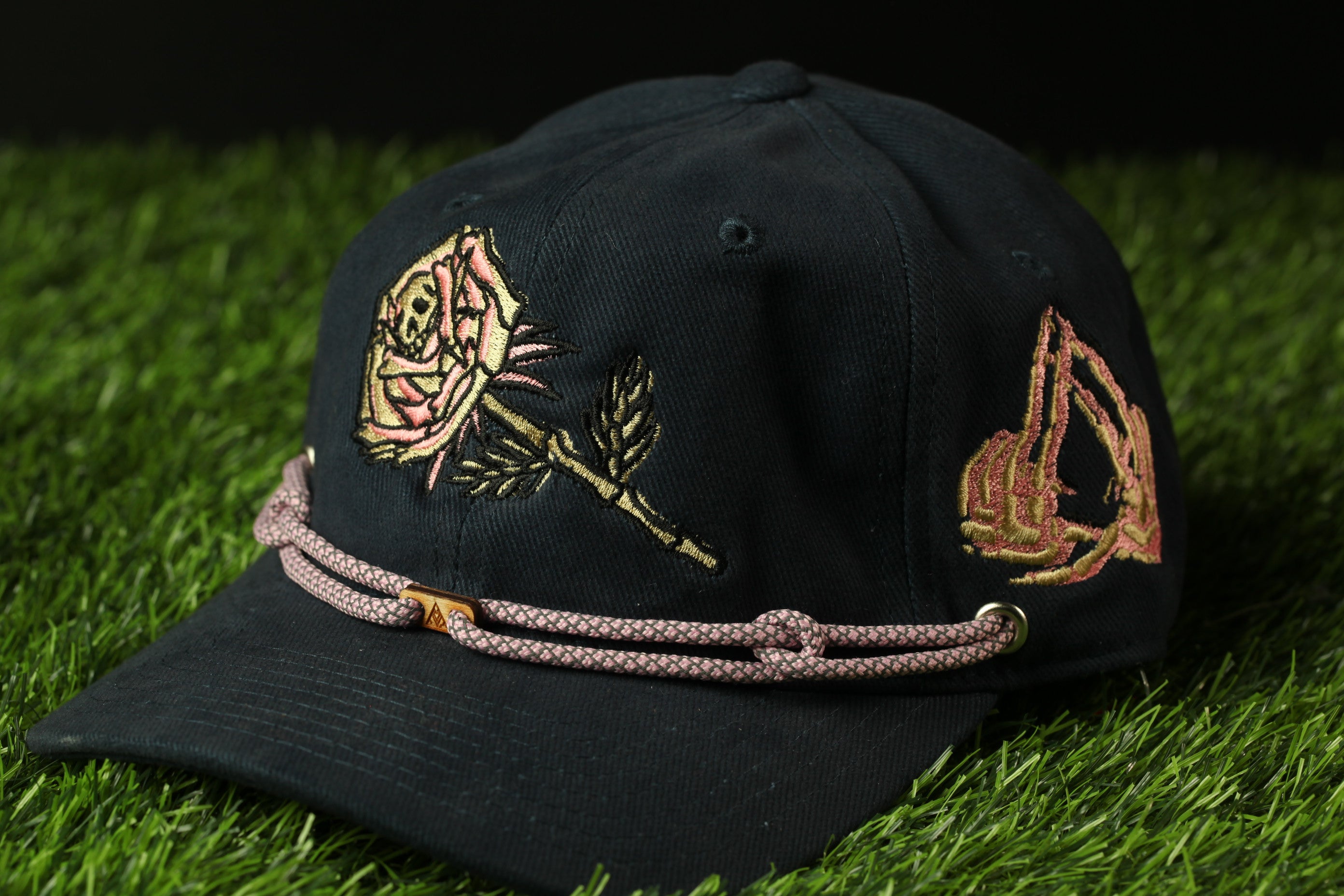 A black baseball cap with gold embroidered designs on the front, including a rose and a geometric pattern, is shown on a grassy surface.
