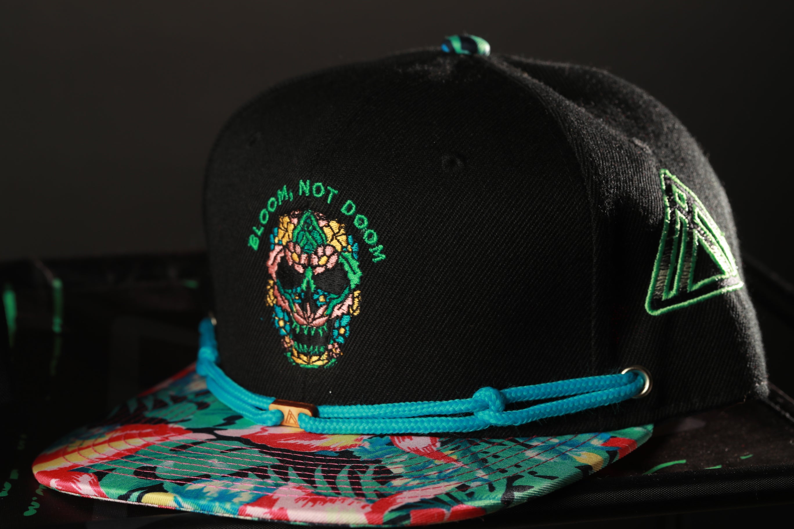 A black baseball cap with a colorful floral pattern and a skull design on the front, along with a green triangle patch on the side.