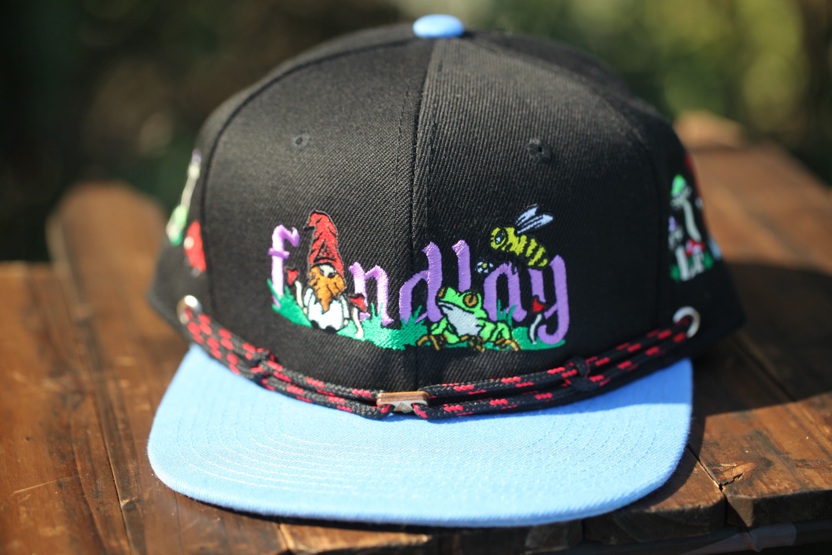 A black baseball cap with colorful embroidered designs, including a gnome and a bee, sits on a wooden surface.