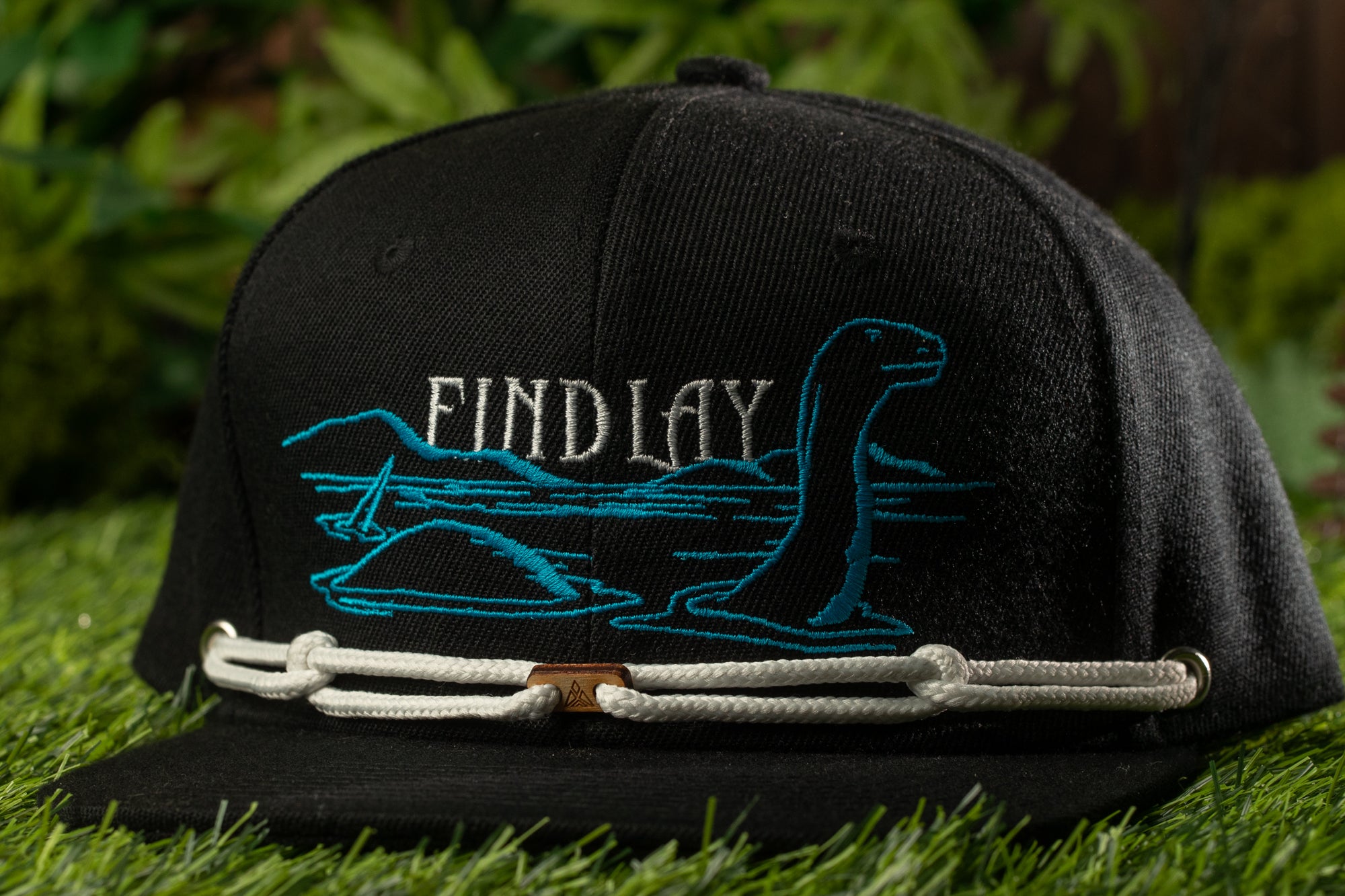 A black baseball cap with a blue and white embroidered design on the front, featuring a large blue dinosaur-like creature and the text "FIND LAY" on the brim.