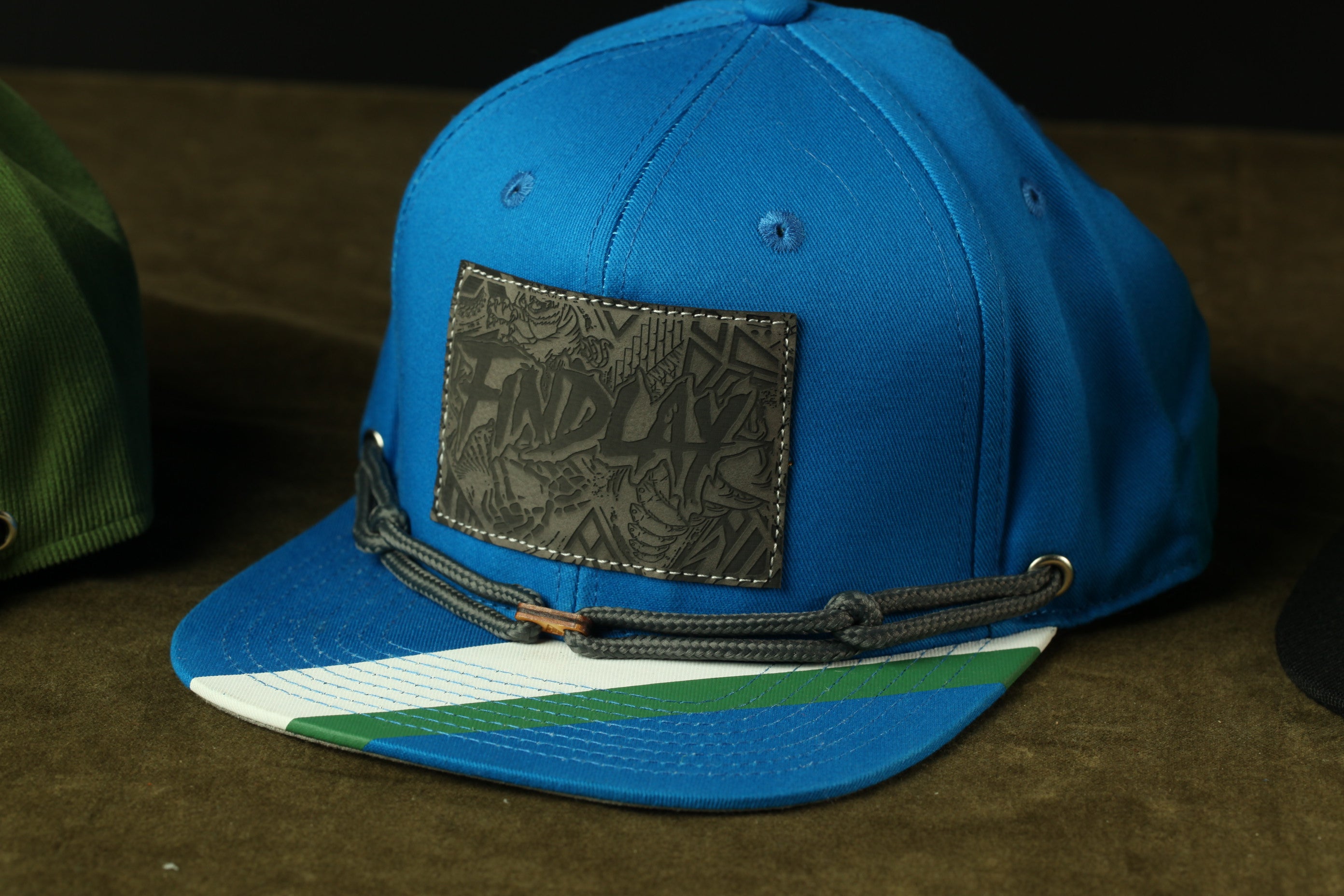 A blue baseball cap with a black patch featuring a graphic design and a green and white striped brim.