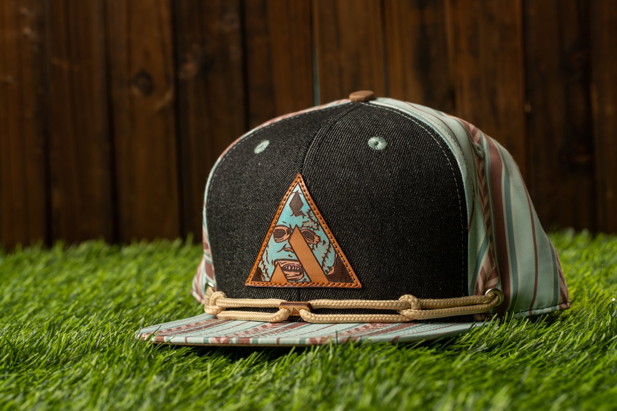 A black baseball cap with a colorful triangle patch featuring a cartoon character is placed on a grassy surface.