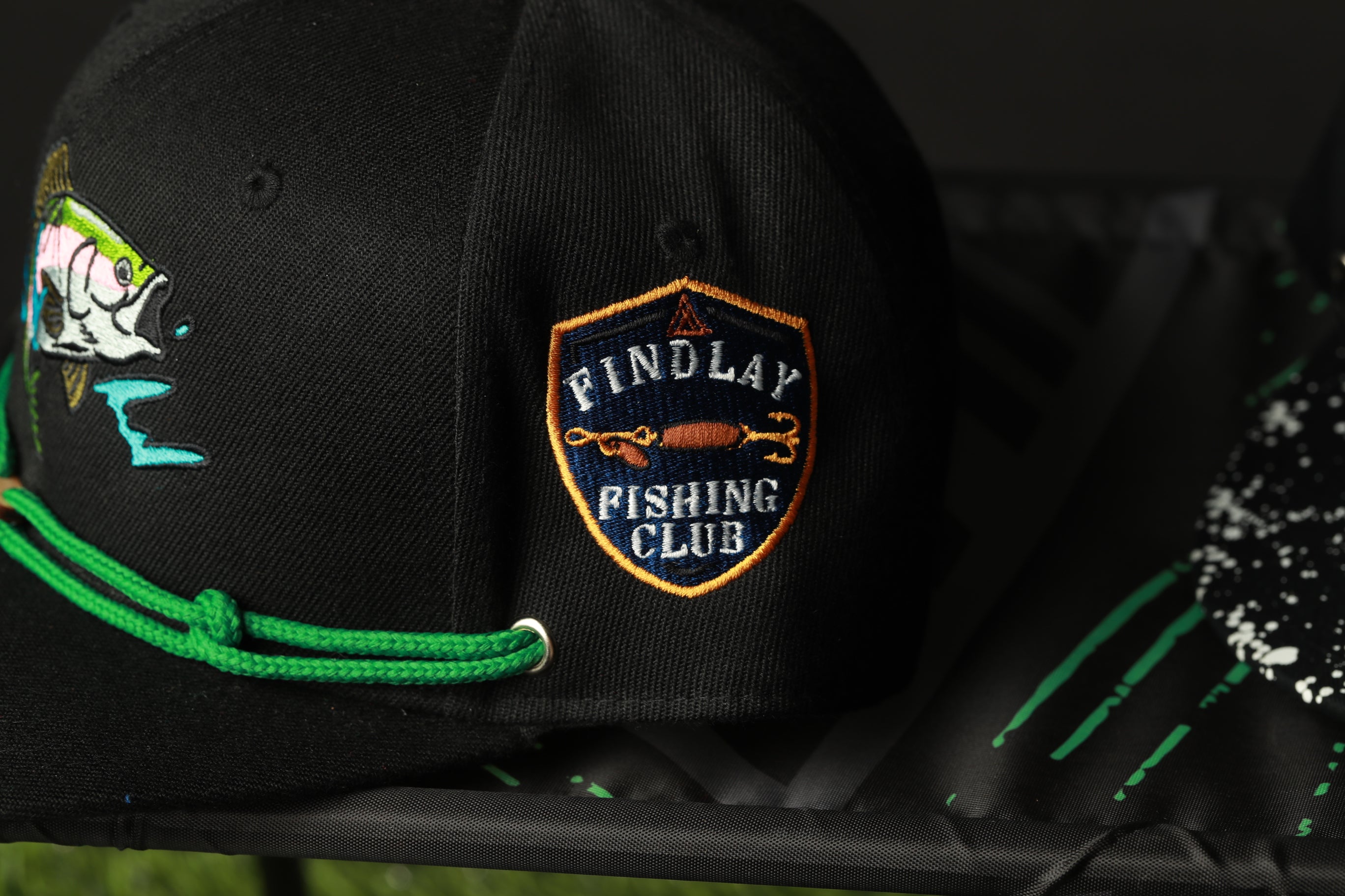 A black baseball cap with a green string and a patch on the front that says "FIND LAY FISHING CLUB".
