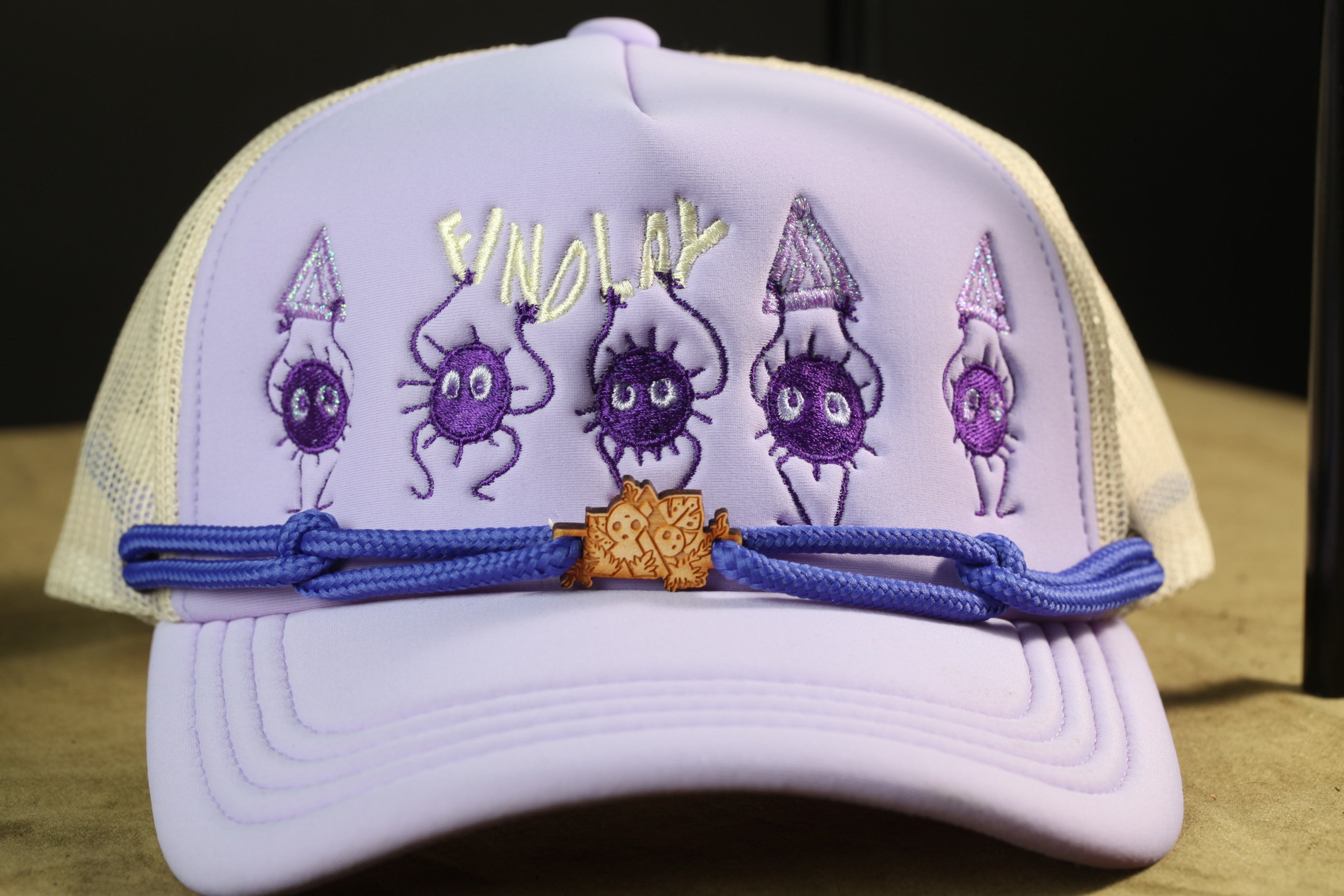 A purple baseball cap with purple and yellow cartoon-like characters on it, and a blue string or strap around the brim.