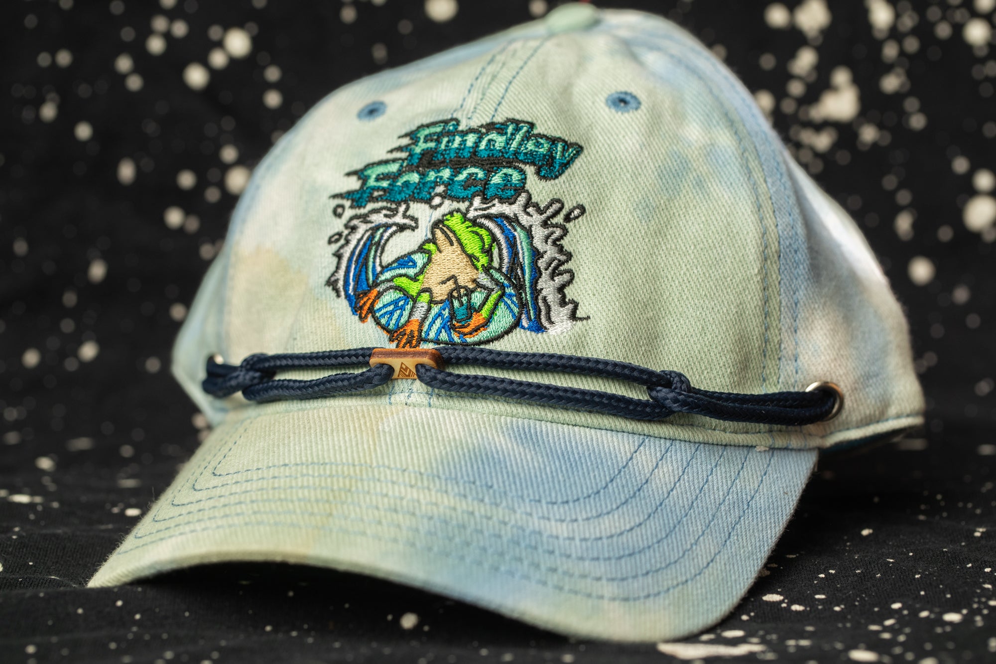 A blue baseball cap with a tie-dye design and a small fish embroidered on it, sitting on a black background with white splatters.