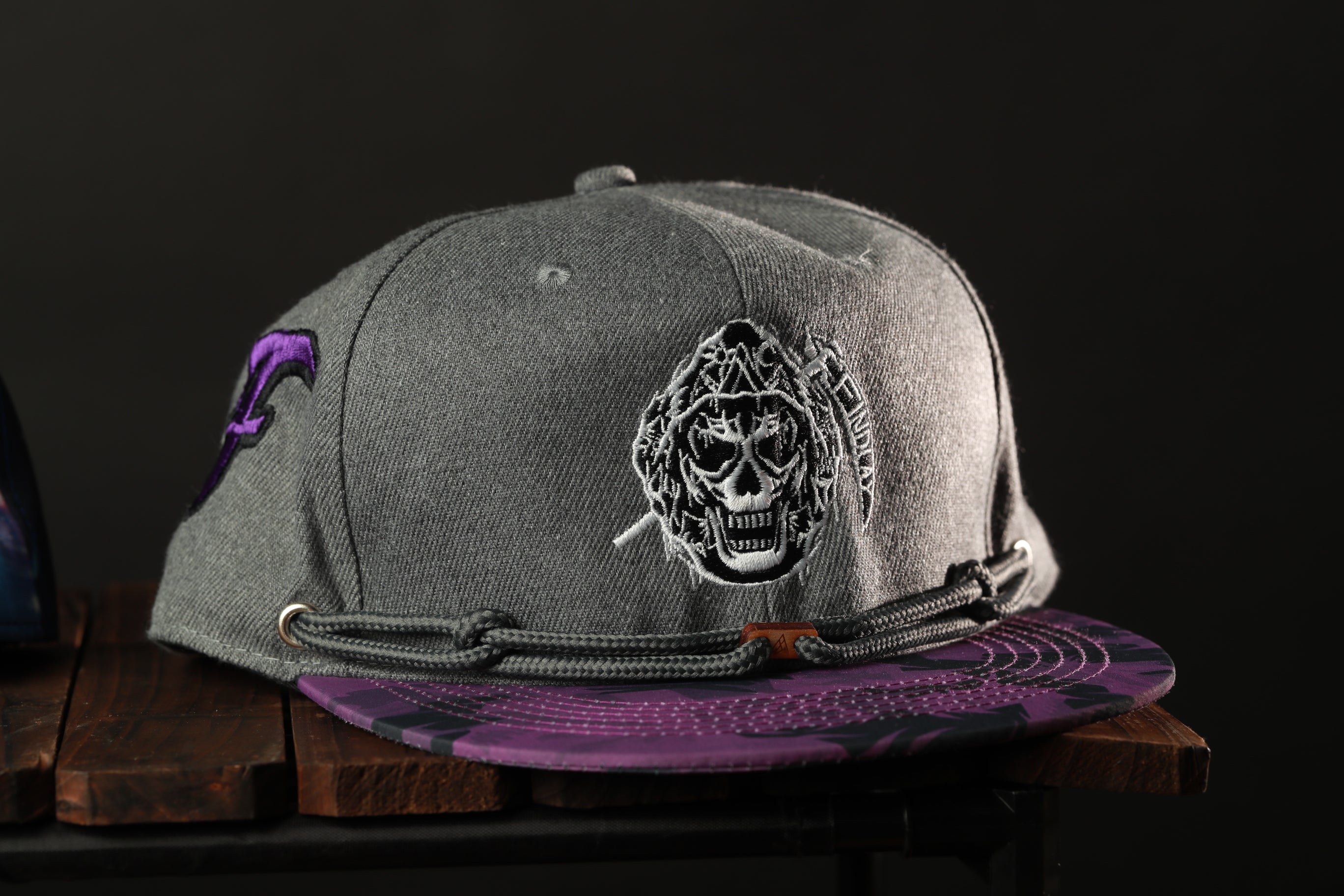 A gray baseball cap with a purple and black patterned brim, featuring a skull design on the front.