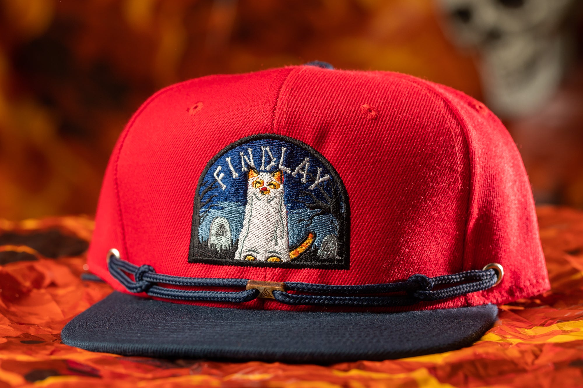 A red baseball cap with a cat embroidered on it, sitting on a red and orange patterned fabric.