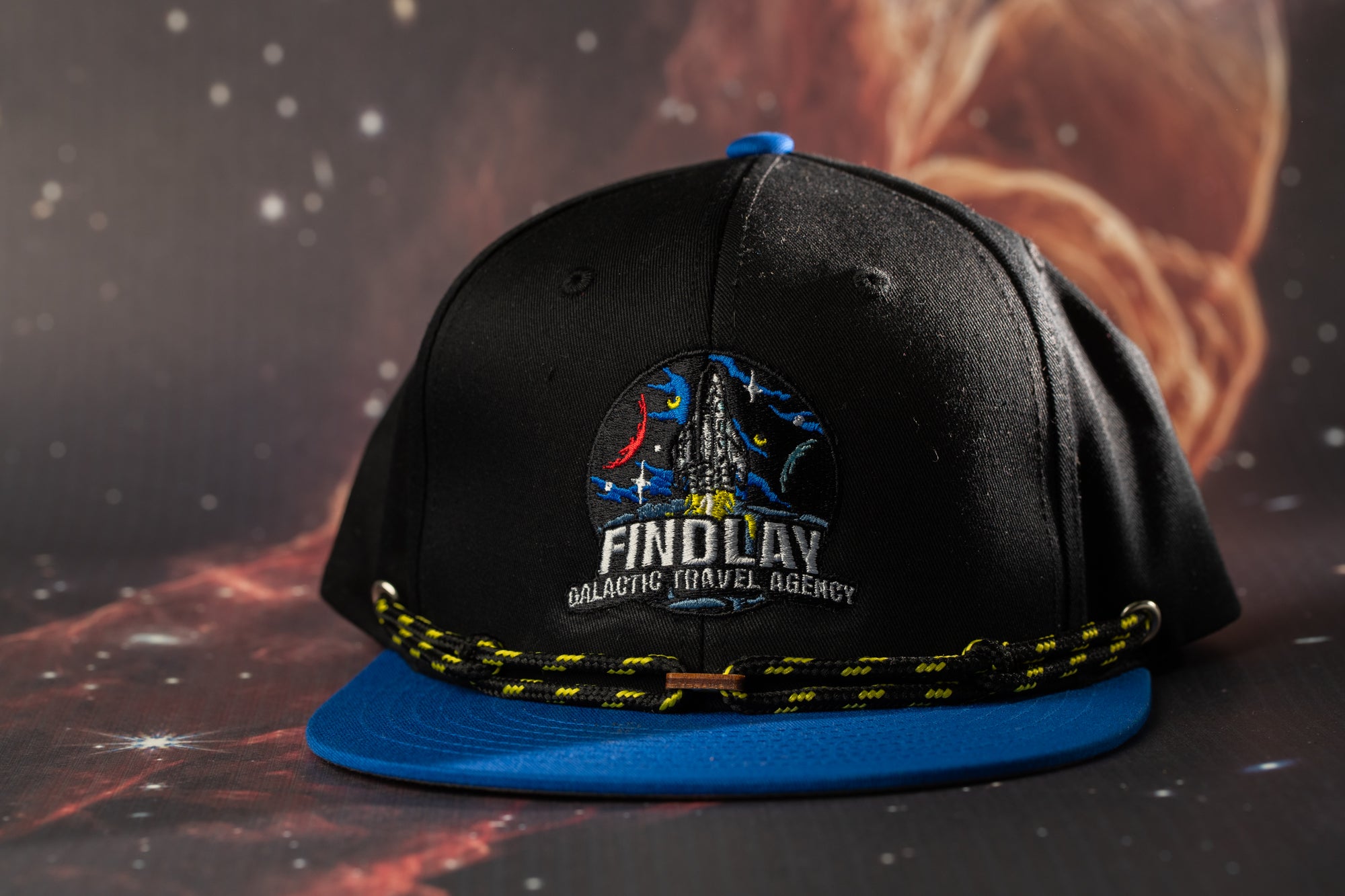 A black baseball cap with a blue brim and a circular logo on the front, featuring the text "FINDOLAY Galactic Travel Agency".