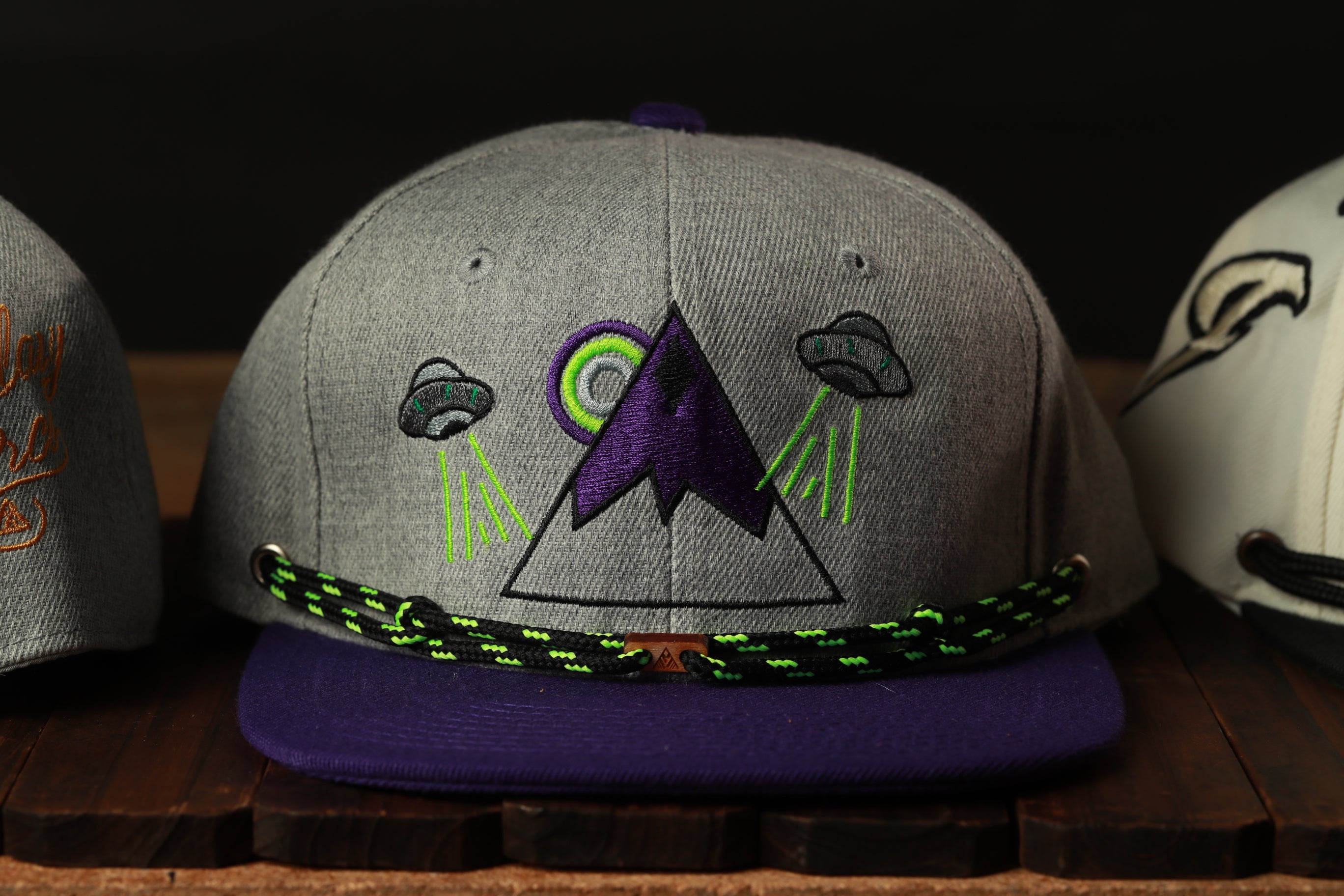 A gray baseball cap with purple and green accents, featuring an embroidered design of a mountain and two alien spacecraft.