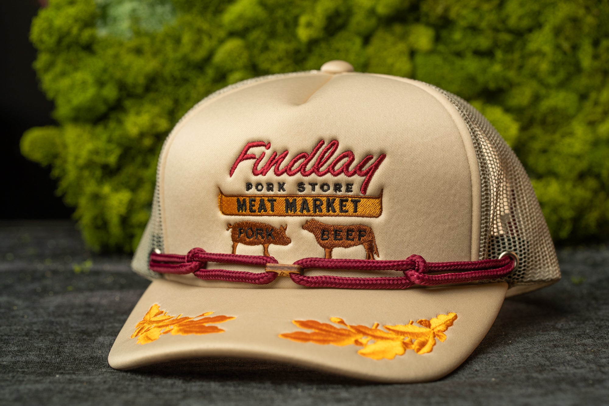 A tan baseball cap with embroidered text and images of pigs, placed on a gray surface against a green background.