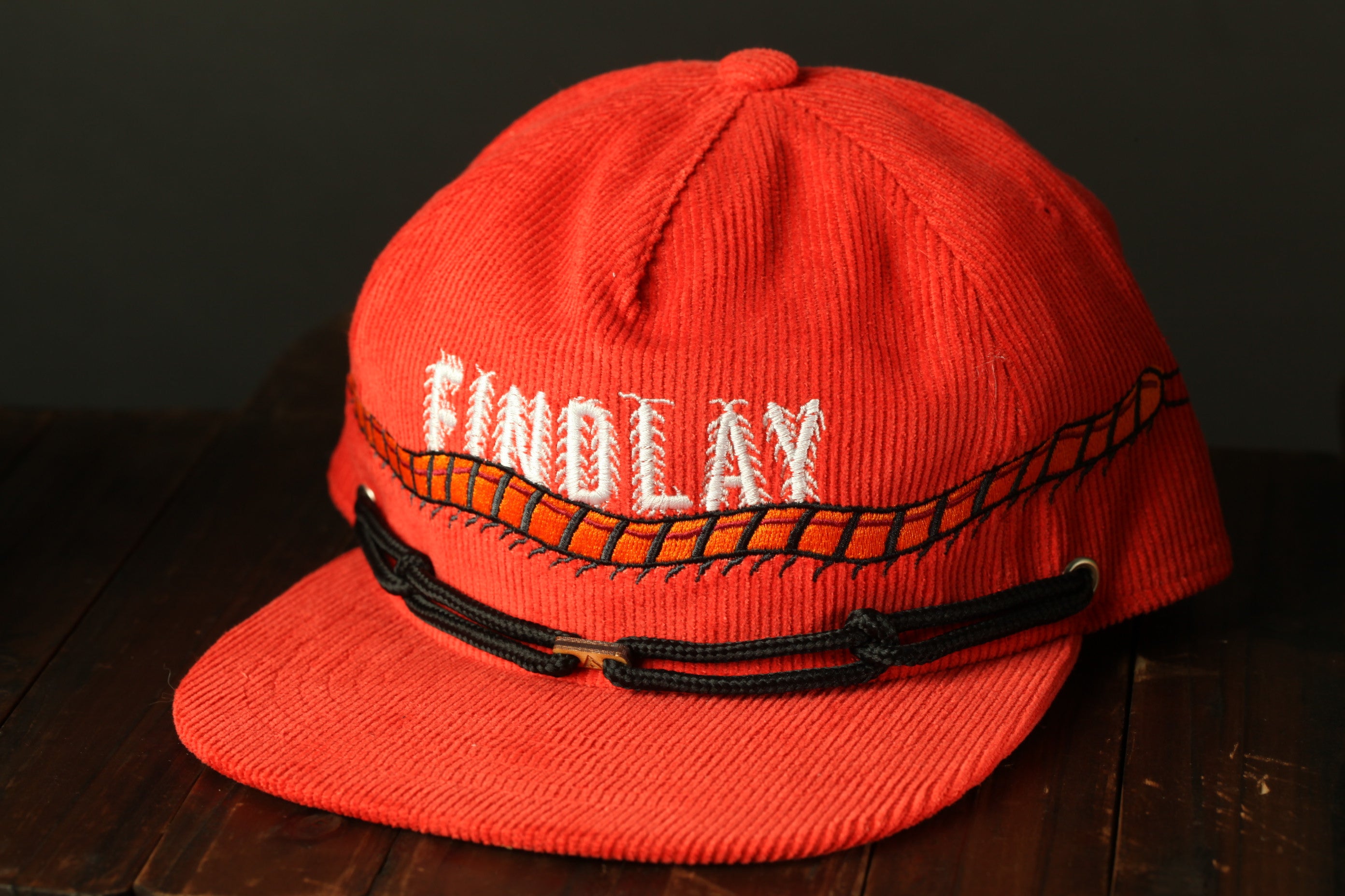 A red baseball cap with the word "FIND LAY" embroidered on it, sitting on a wooden surface.
