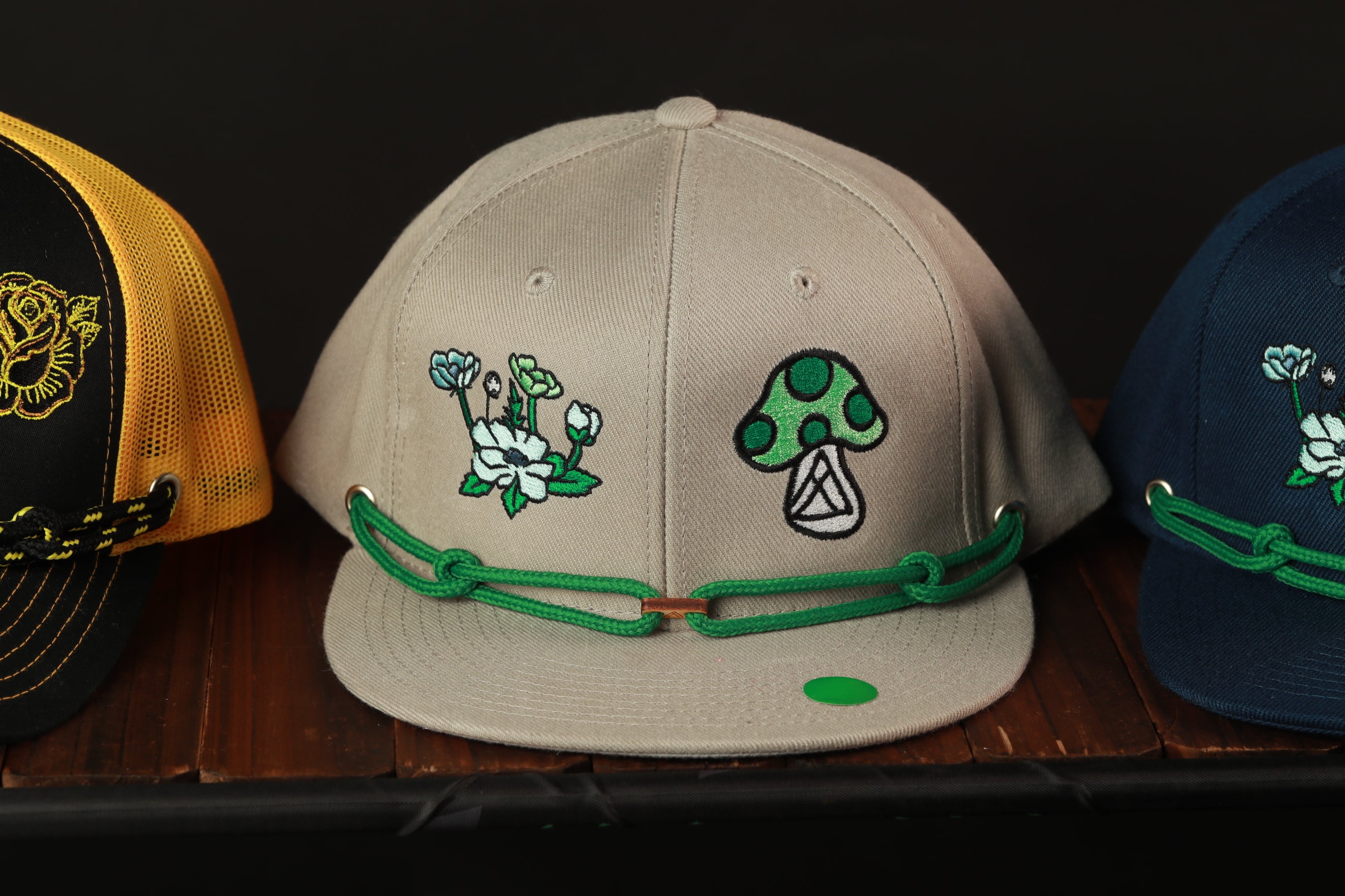 Three baseball caps with different designs on them, including flowers and mushrooms, are displayed on a wooden surface.