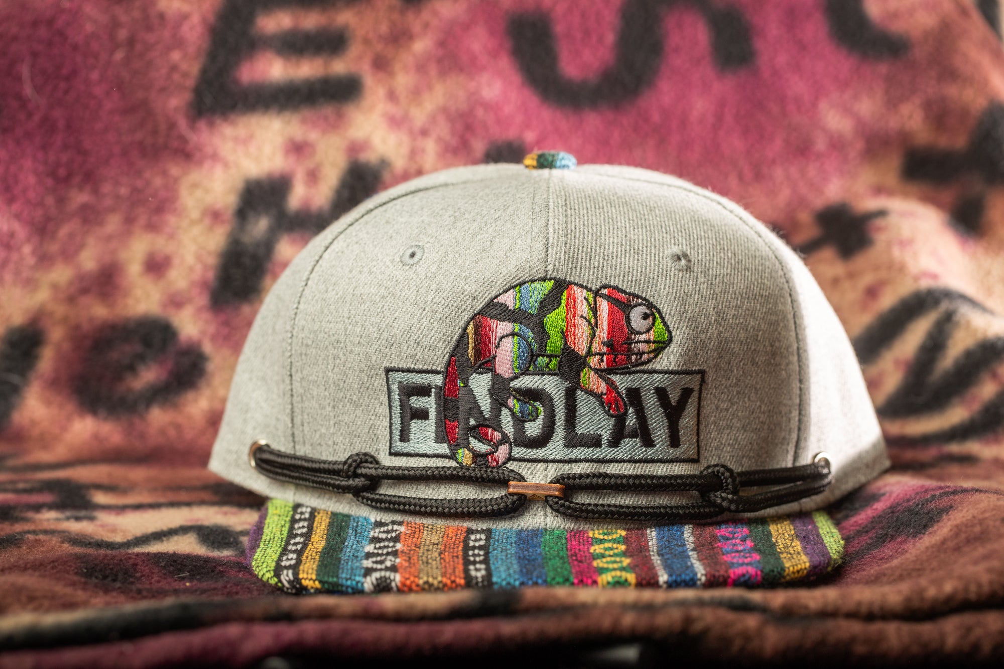 A gray baseball cap with a colorful chameleon design and the word "FIND PLAY" printed on it, placed on a multicolored blanket.
