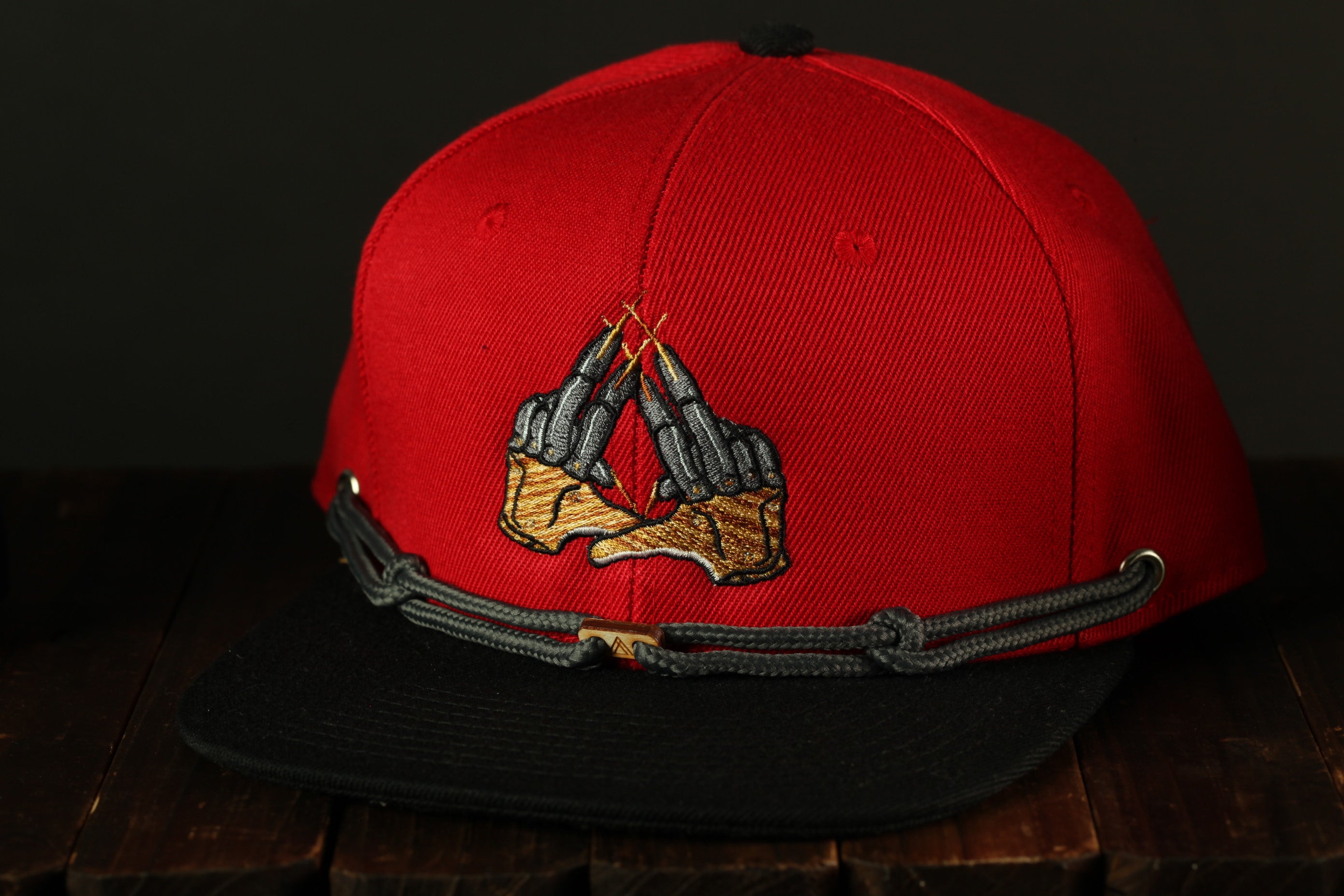 A red baseball cap with a black brim and a black band around the front, featuring an embroidered design of two hands.