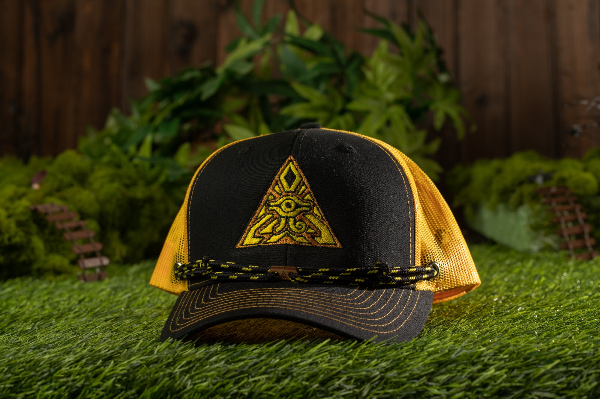 A black baseball cap with a yellow triangle design and a black strap, placed on a grassy surface with a wooden fence and greenery in the background.