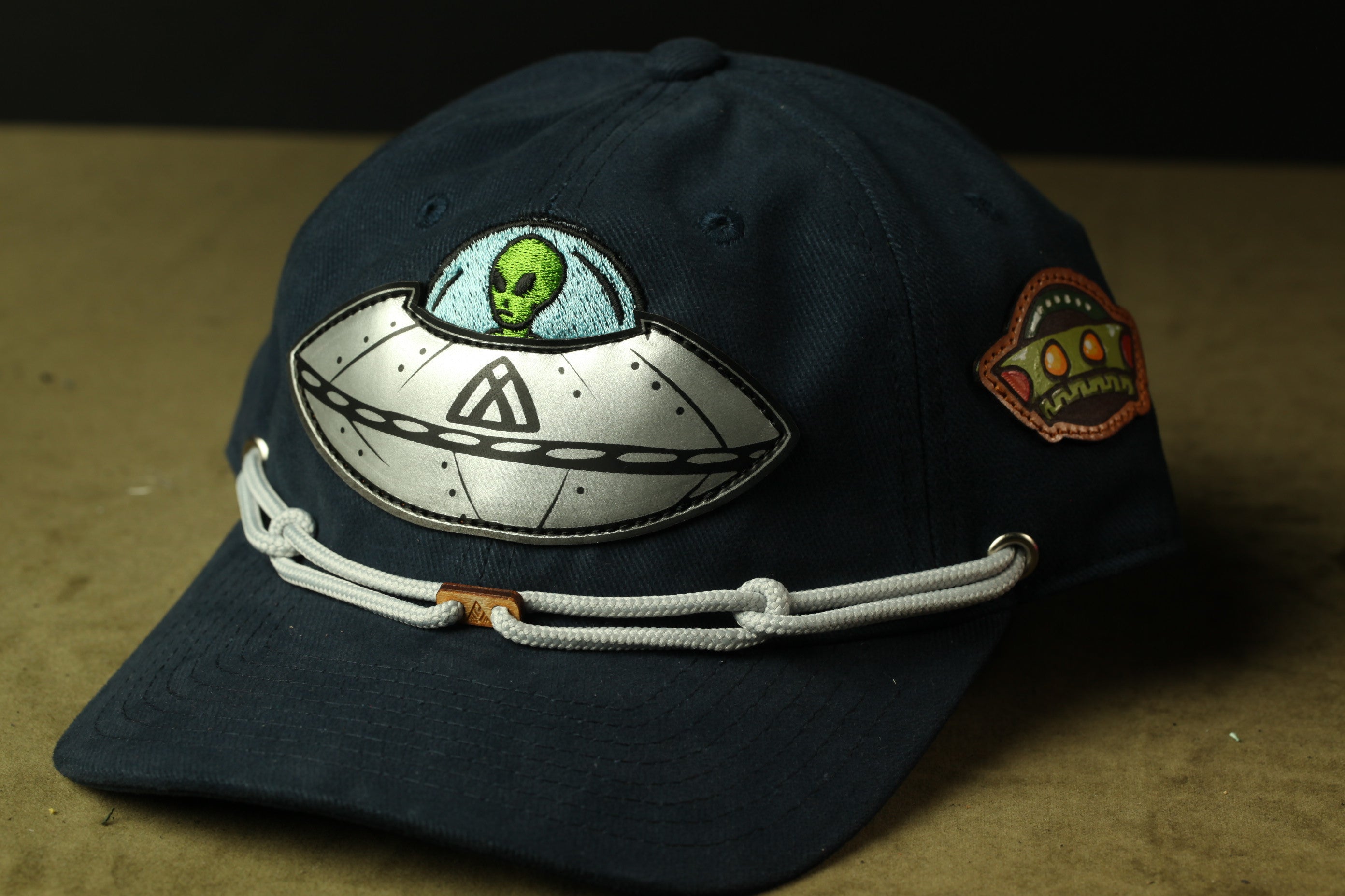 A dark blue baseball cap with a unique alien design on the front, featuring a green alien face and a spaceship motif.