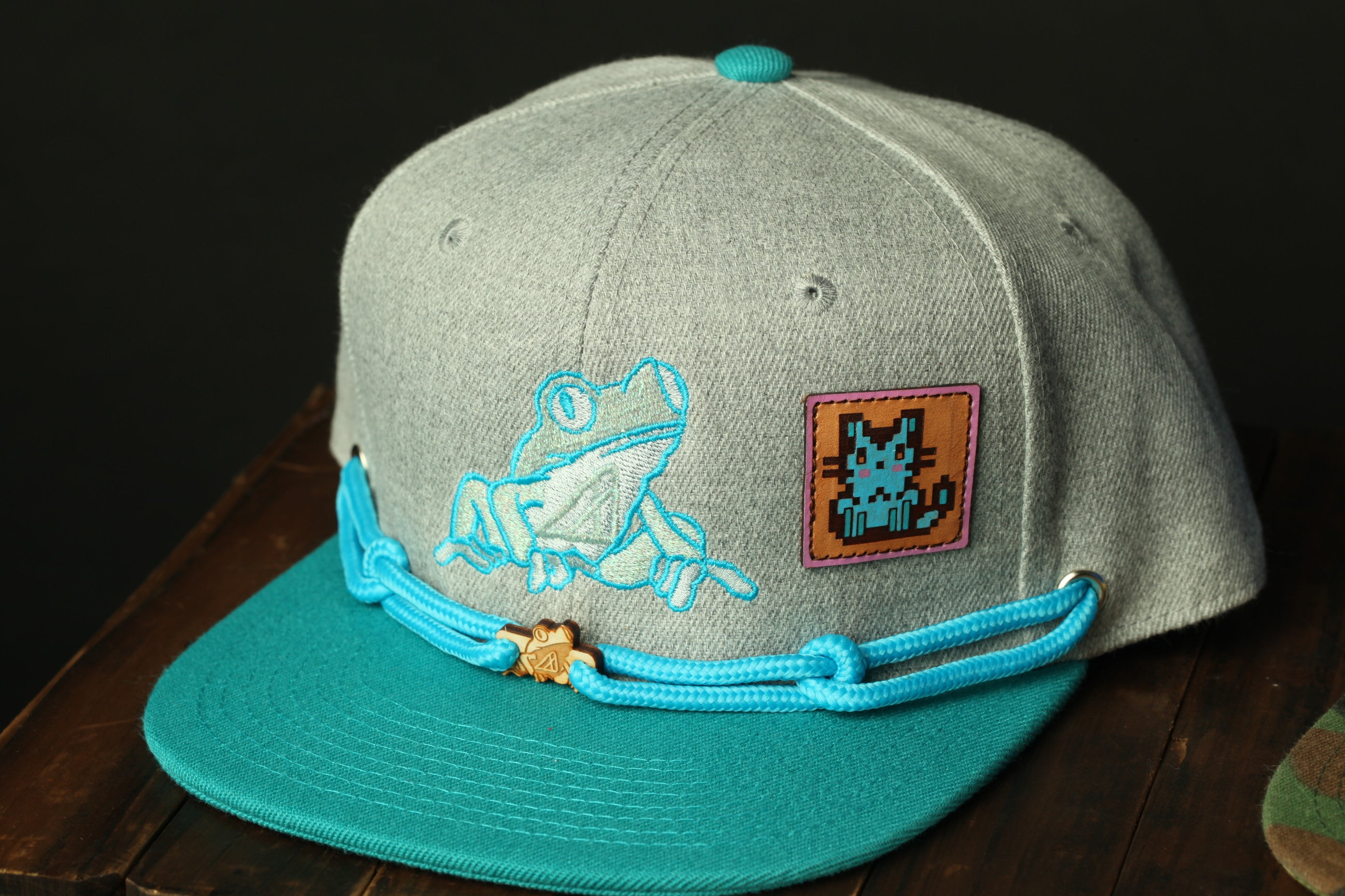 A gray baseball cap with a blue and teal color scheme, featuring a frog embroidered on the front and a cat embroidered on the side.