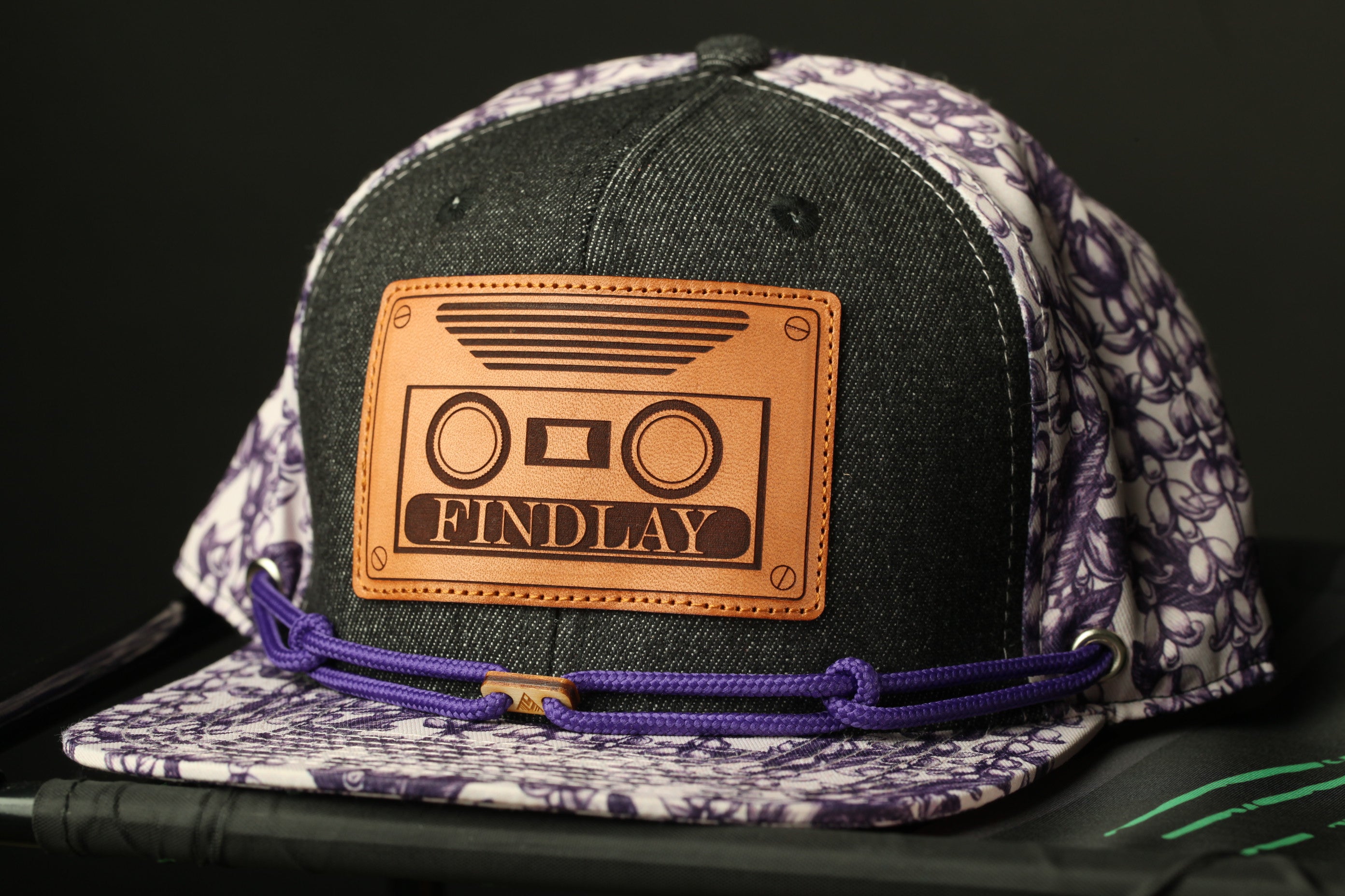 A black and purple baseball cap with a leather patch that says "FIND LAY" on it.