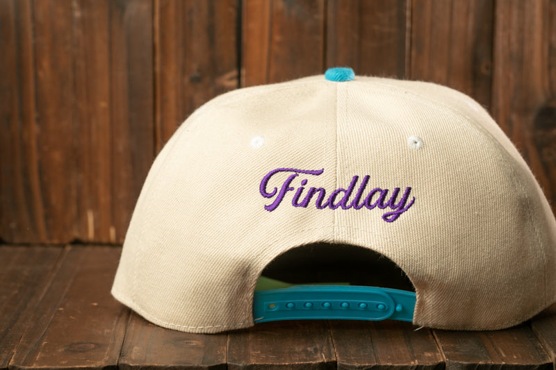 A beige baseball cap with the word "Findlay" embroidered on it, sitting on a wooden surface.