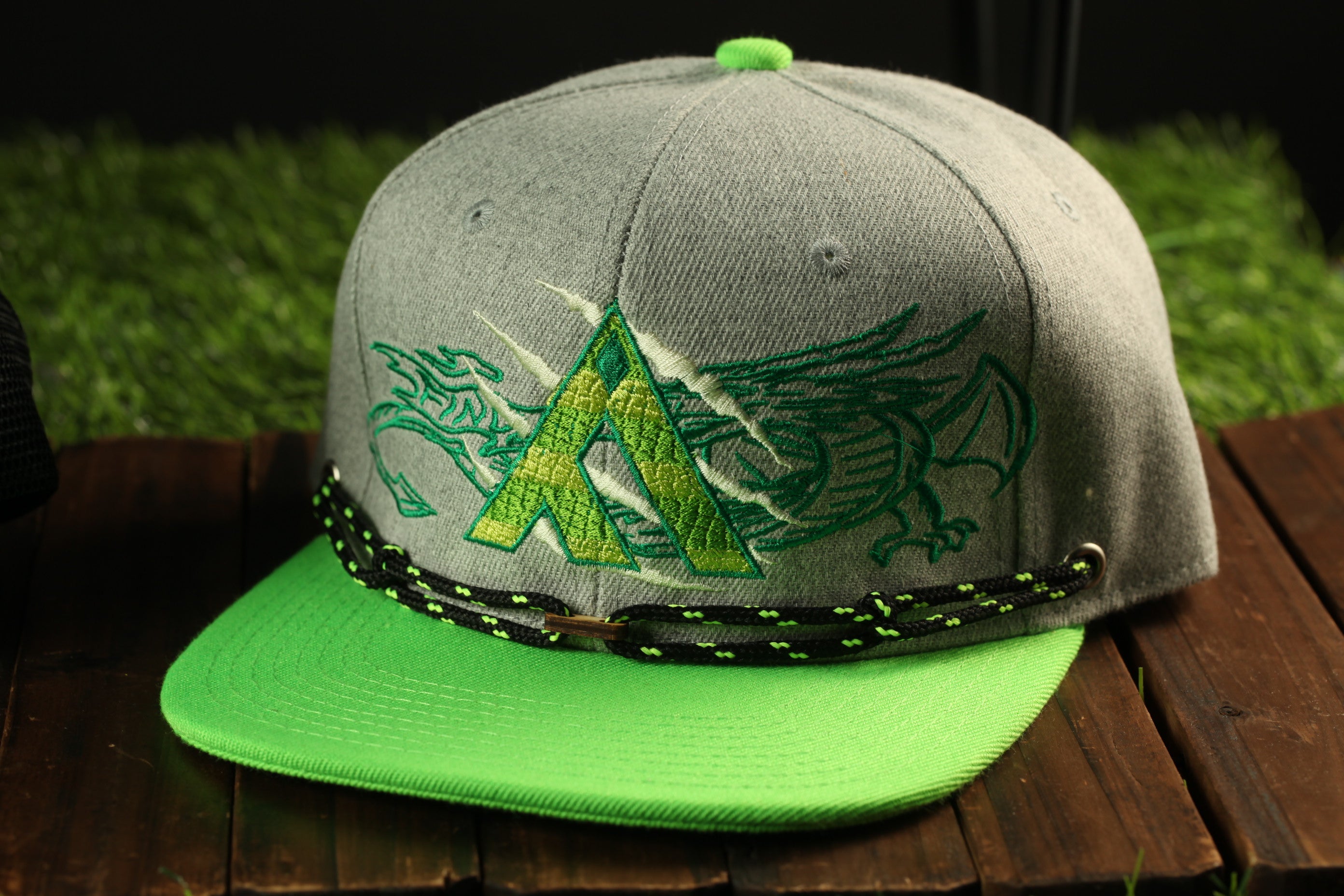 A gray baseball cap with a green brim and a dragon design on the front, sitting on a wooden surface.