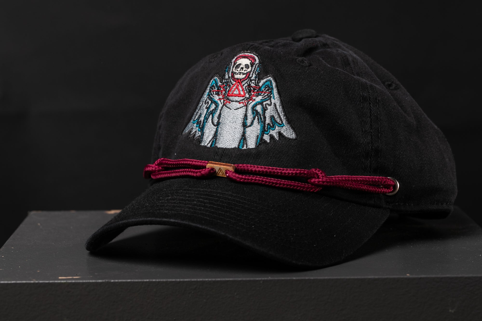 A black baseball cap with a skull and crossbones design on the front, and a red string or strap around the brim.