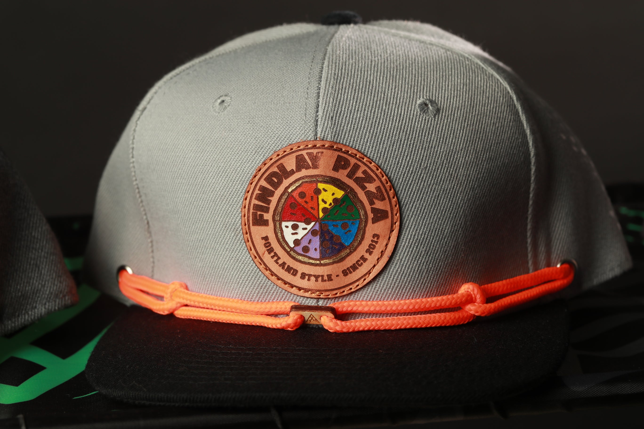 A gray baseball cap with a circular patch on the front, featuring a colorful design and the text "FINDlay PIZZA" printed on it.