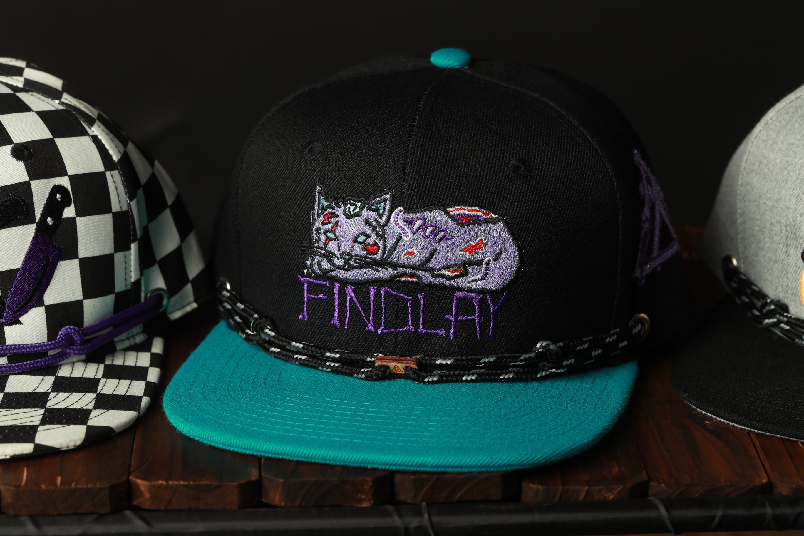 A black baseball cap with a purple cat embroidered on it, sitting on a wooden surface.