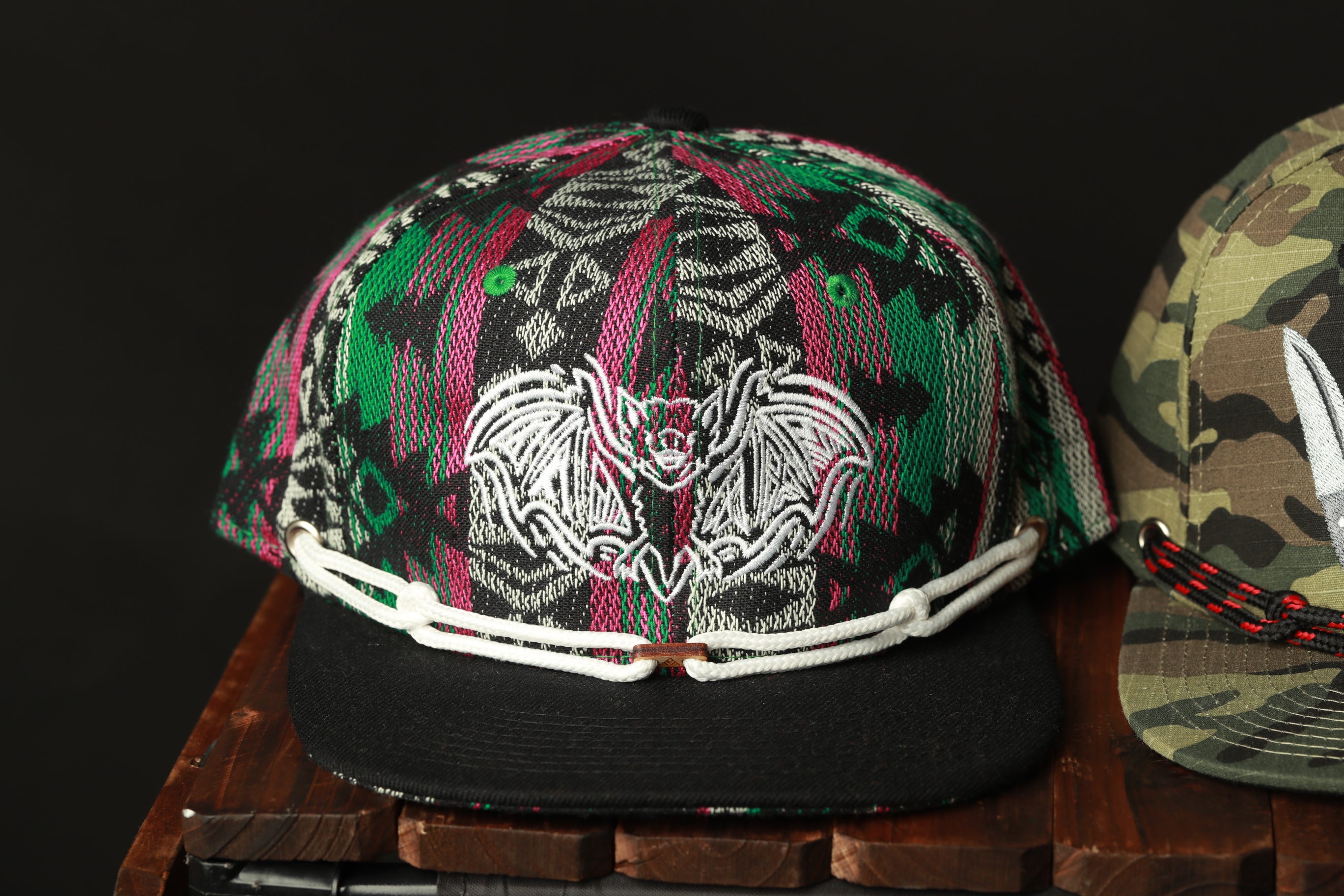 A colorful, patterned baseball cap with a bat design on the front, displayed on a wooden surface next to another cap.