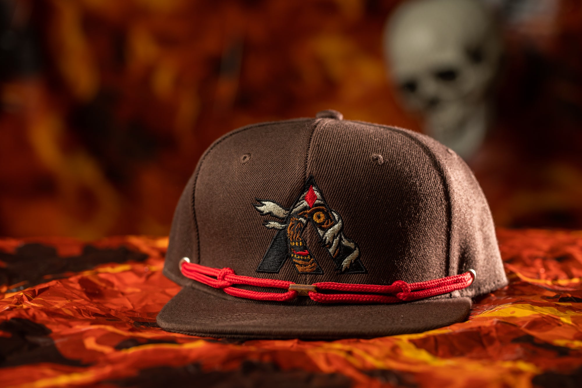 A brown baseball cap with a red string and a graphic design on the front, placed on a red and orange patterned surface.