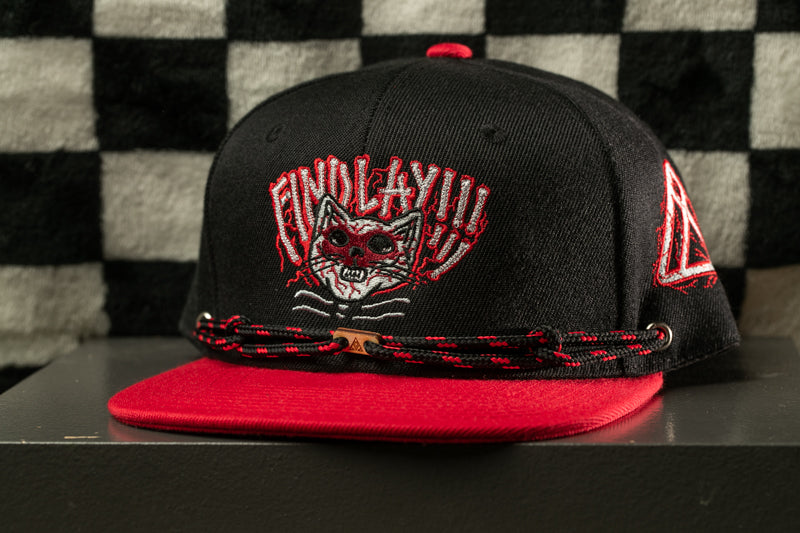A black baseball cap with a red brim and a cat-like design on the front, sitting on a black surface against a checkered background.