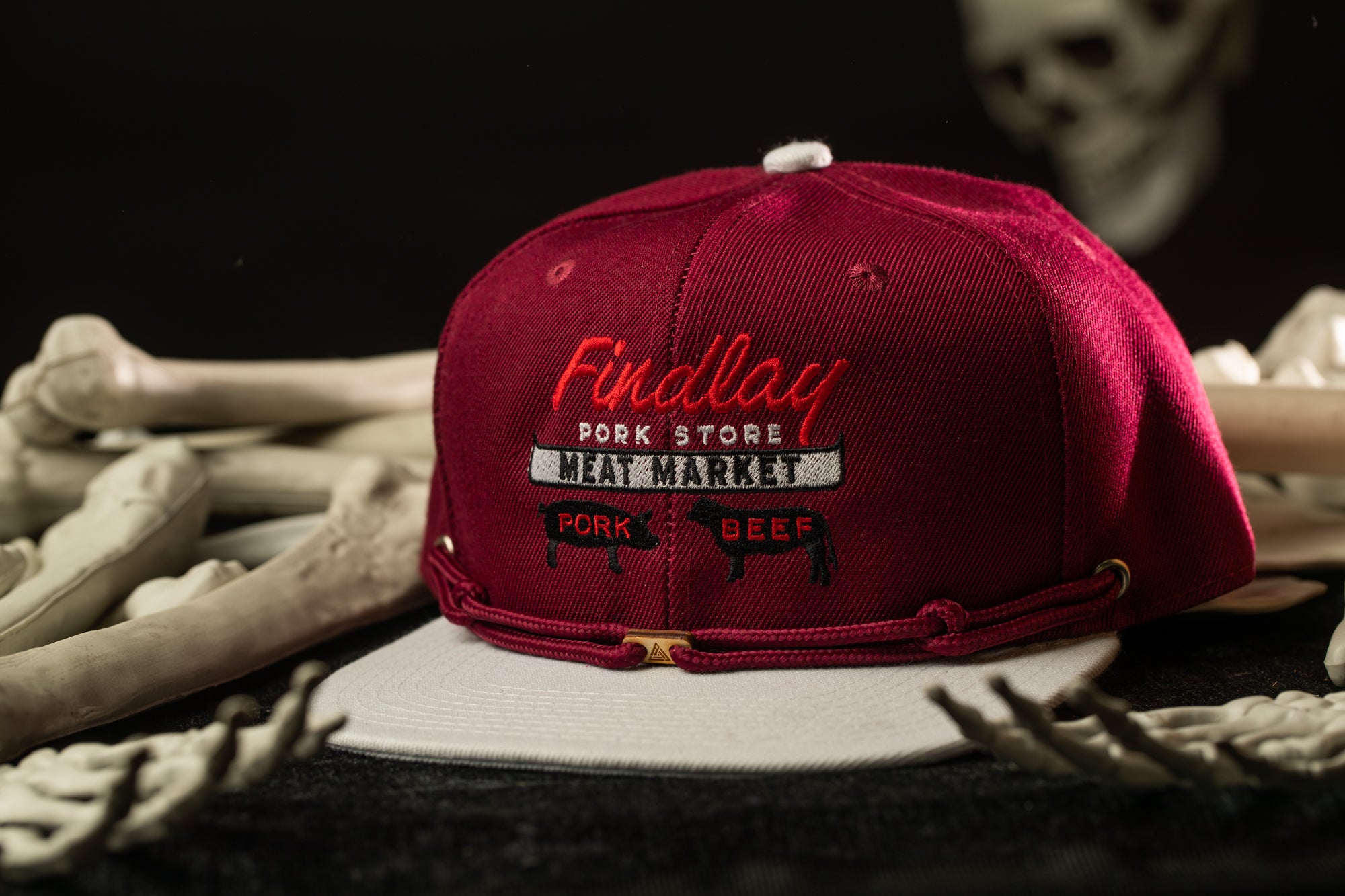 A maroon baseball cap with the text "Findlay Pork Store Meat Market" printed on it, placed on a surface with bones and a skeleton head in the background.