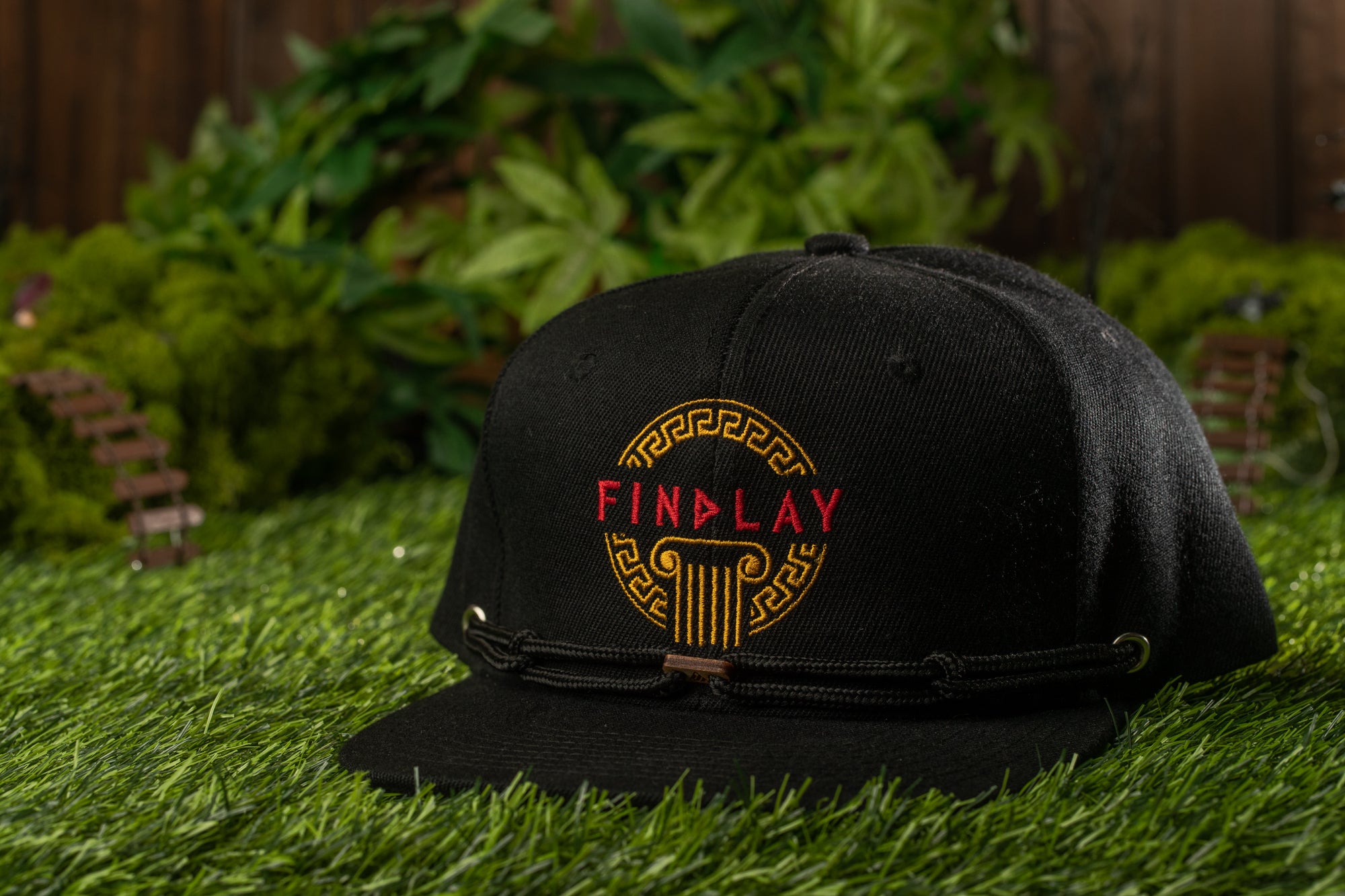 A black baseball cap with the word "FINLAND" printed on it, placed on a grassy surface with a miniature garden in the background.