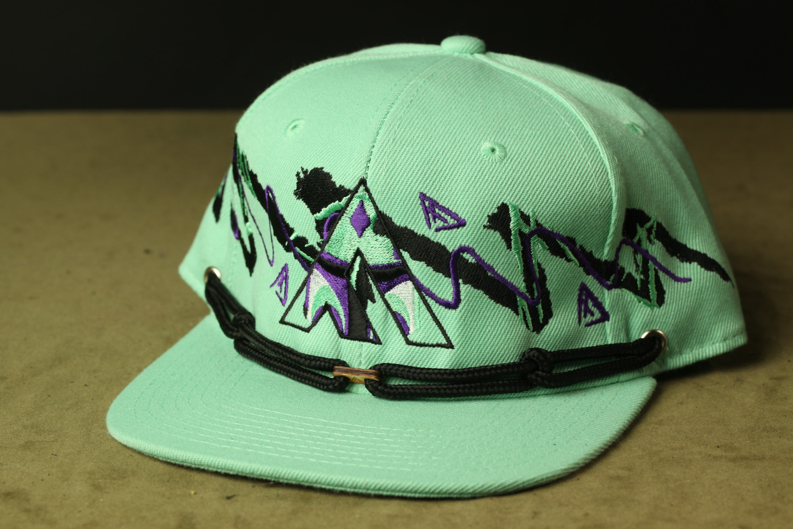 A mint green baseball cap with a black and purple geometric design on the front.
