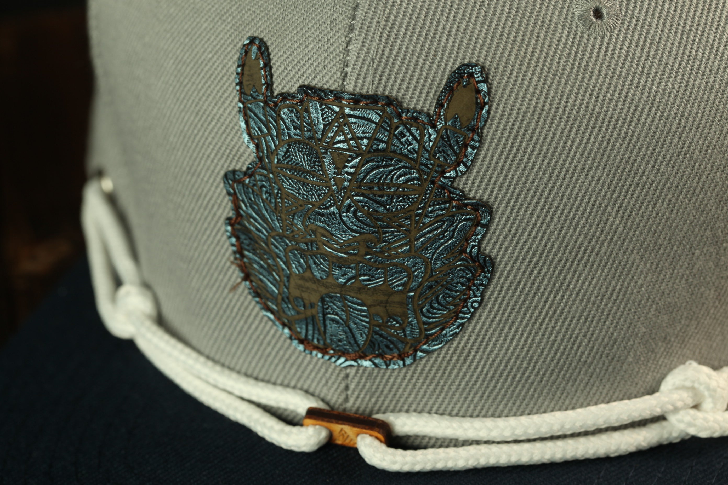 A close-up view of a gray baseball cap with a decorative blue and gold embroidered patch on the front.